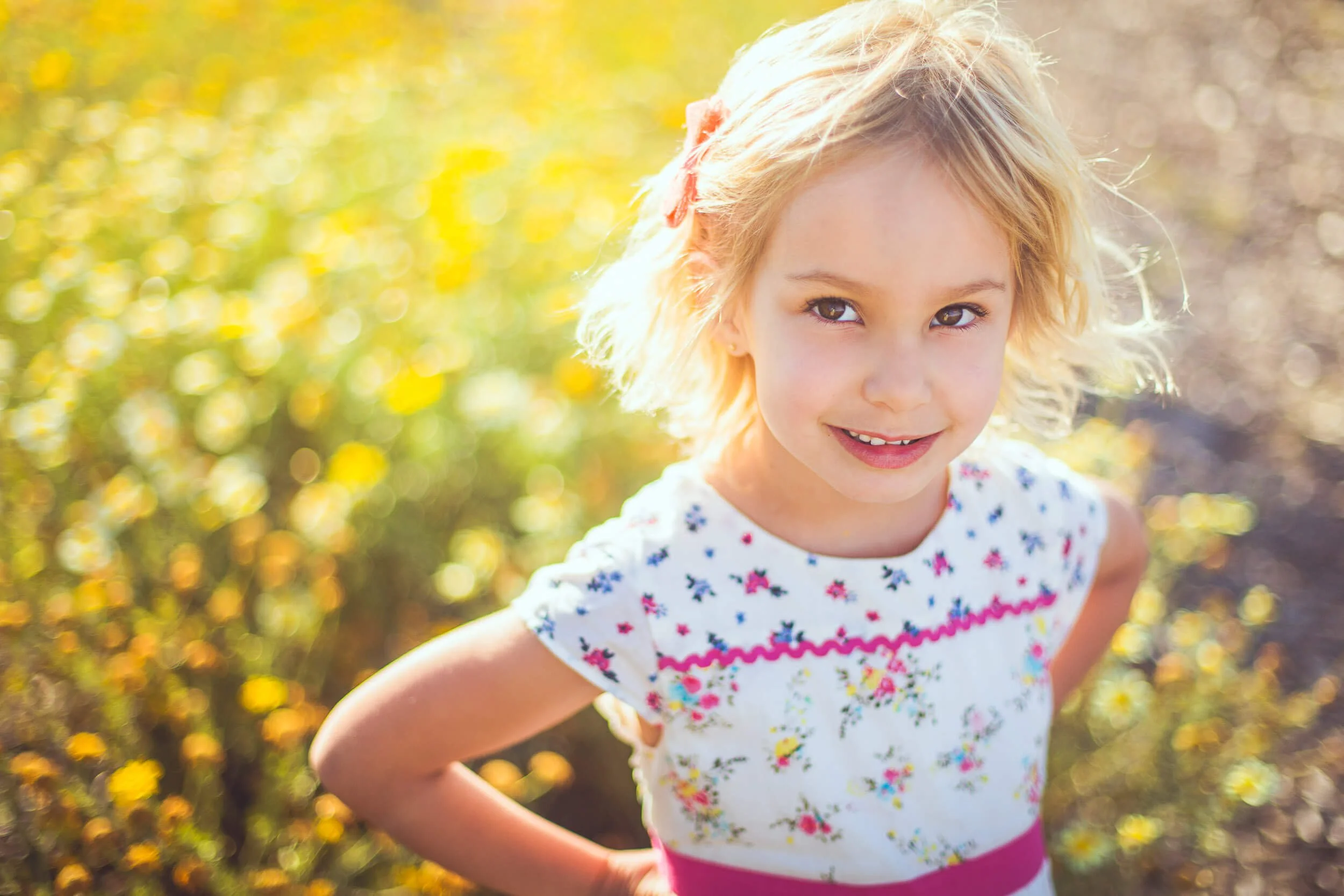 A young girl with blonde curly hair and a pink hair clip smiling outdoors in bright sunlight, wearing a white dress with floral patterns.