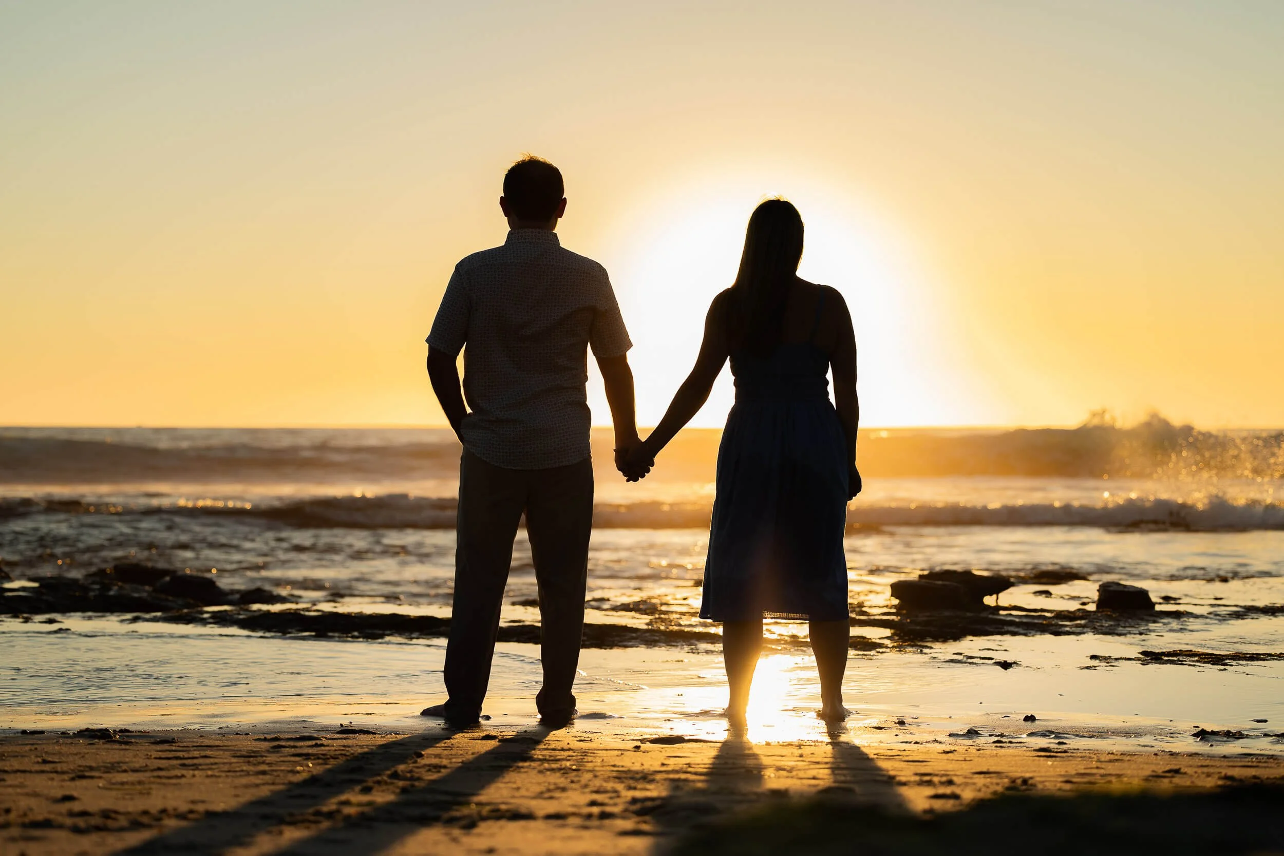 Silhouette of a man and woman holding hands on the beach at sunset, facing the ocean with waves and rocks in the background.