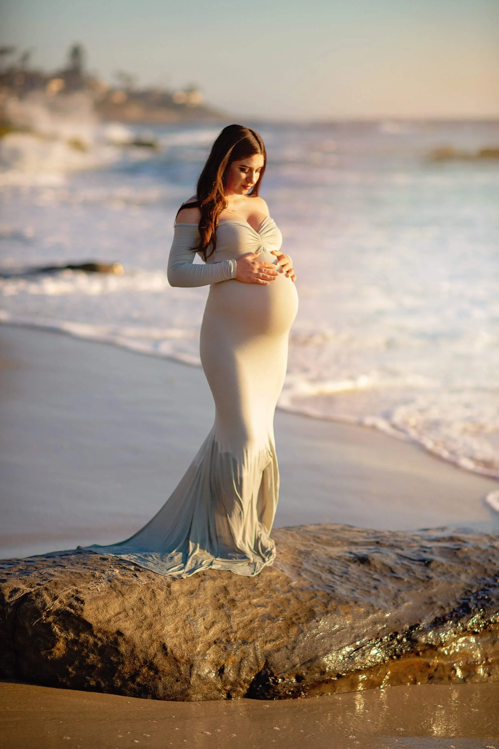 A pregnant woman in an off-shoulder dress standing on a rock on the beach, looking down and holding her belly, with ocean waves in the background.