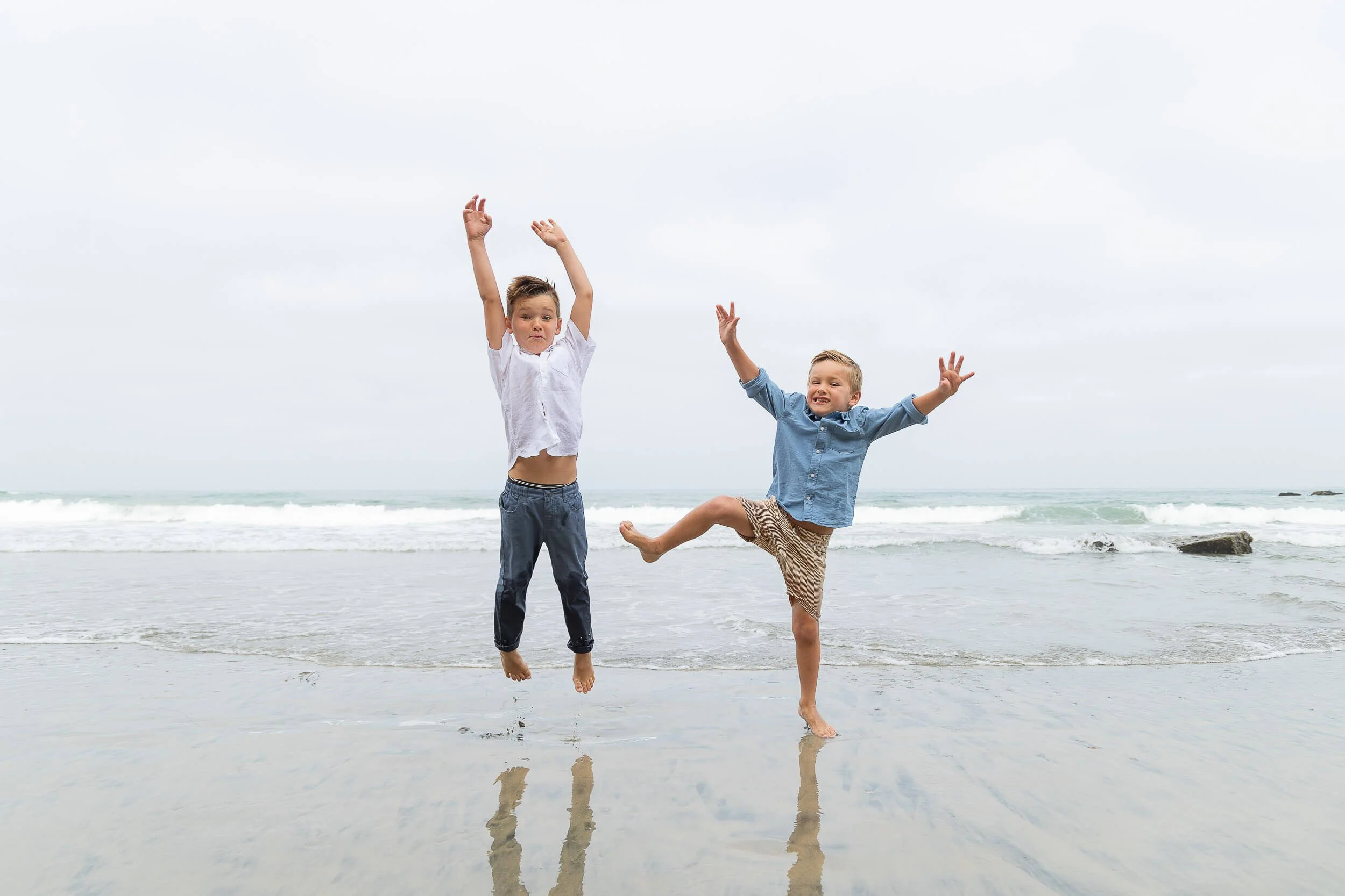 Two young boys playing and jumping on the wet sand by the ocean on a cloudy day.