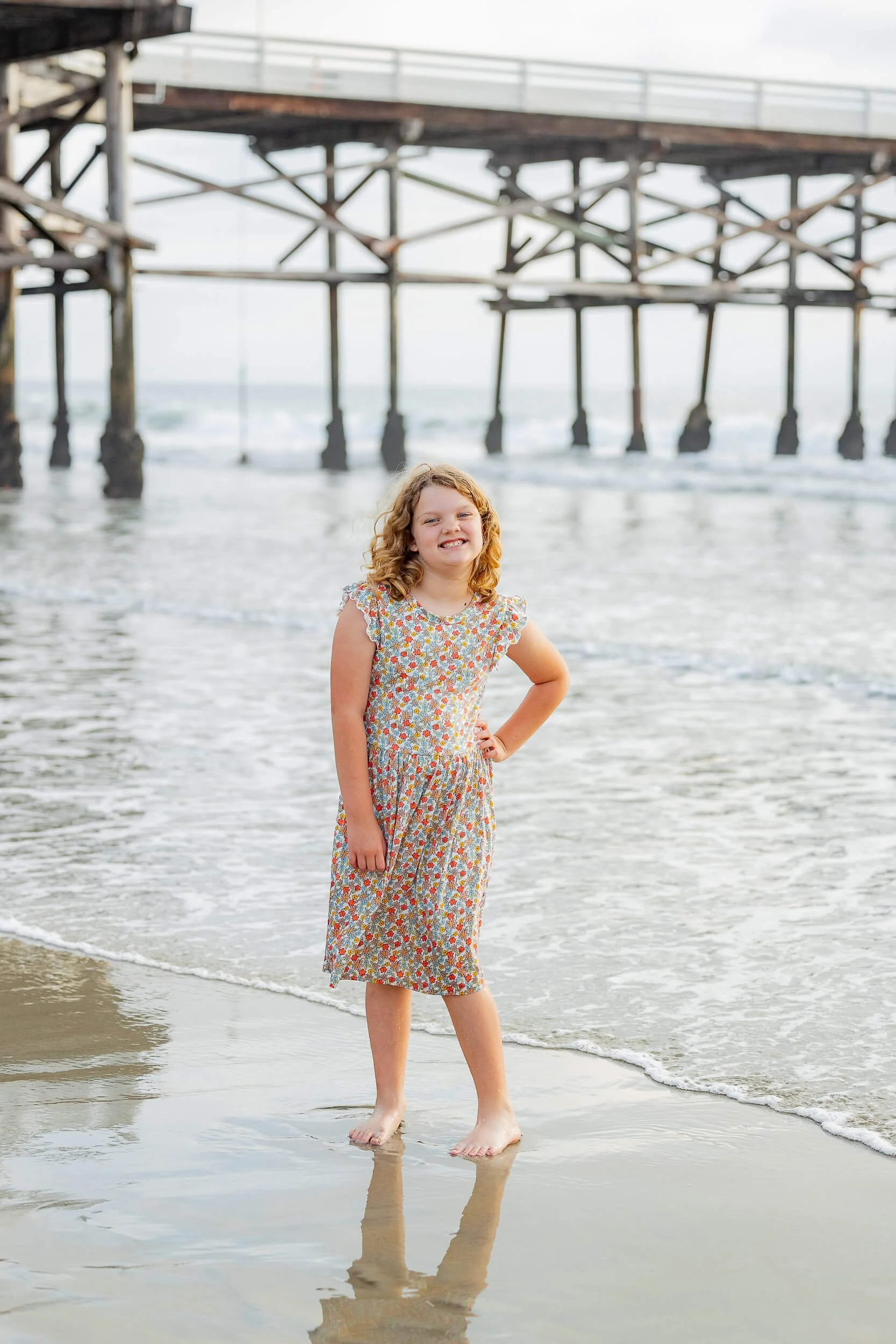 A young girl in a floral dress standing on the beach near the water, with a pier in the background.