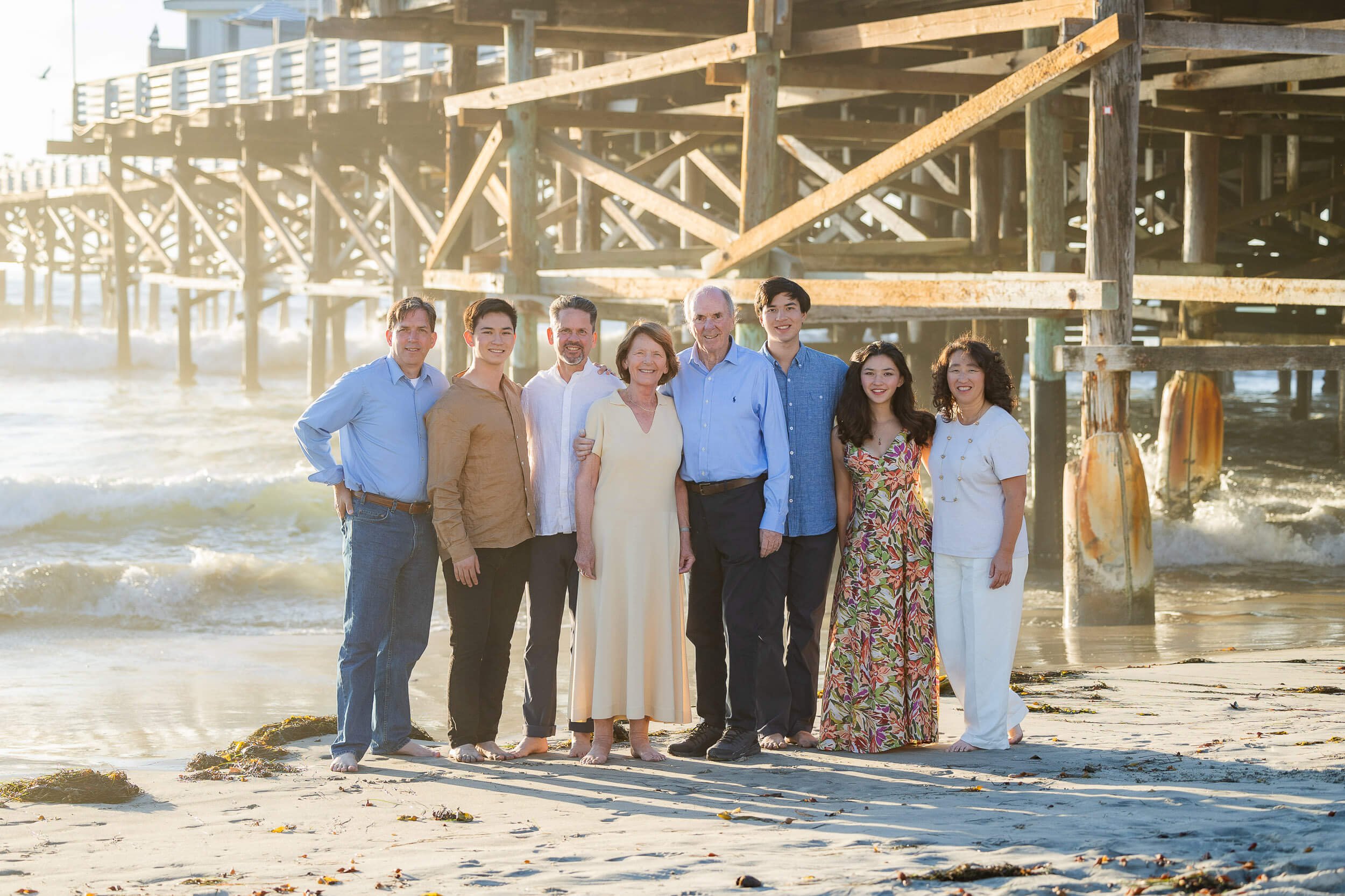 A group of nine people standing on the beach in front of a wooden pier, smiling at the camera during sunset.