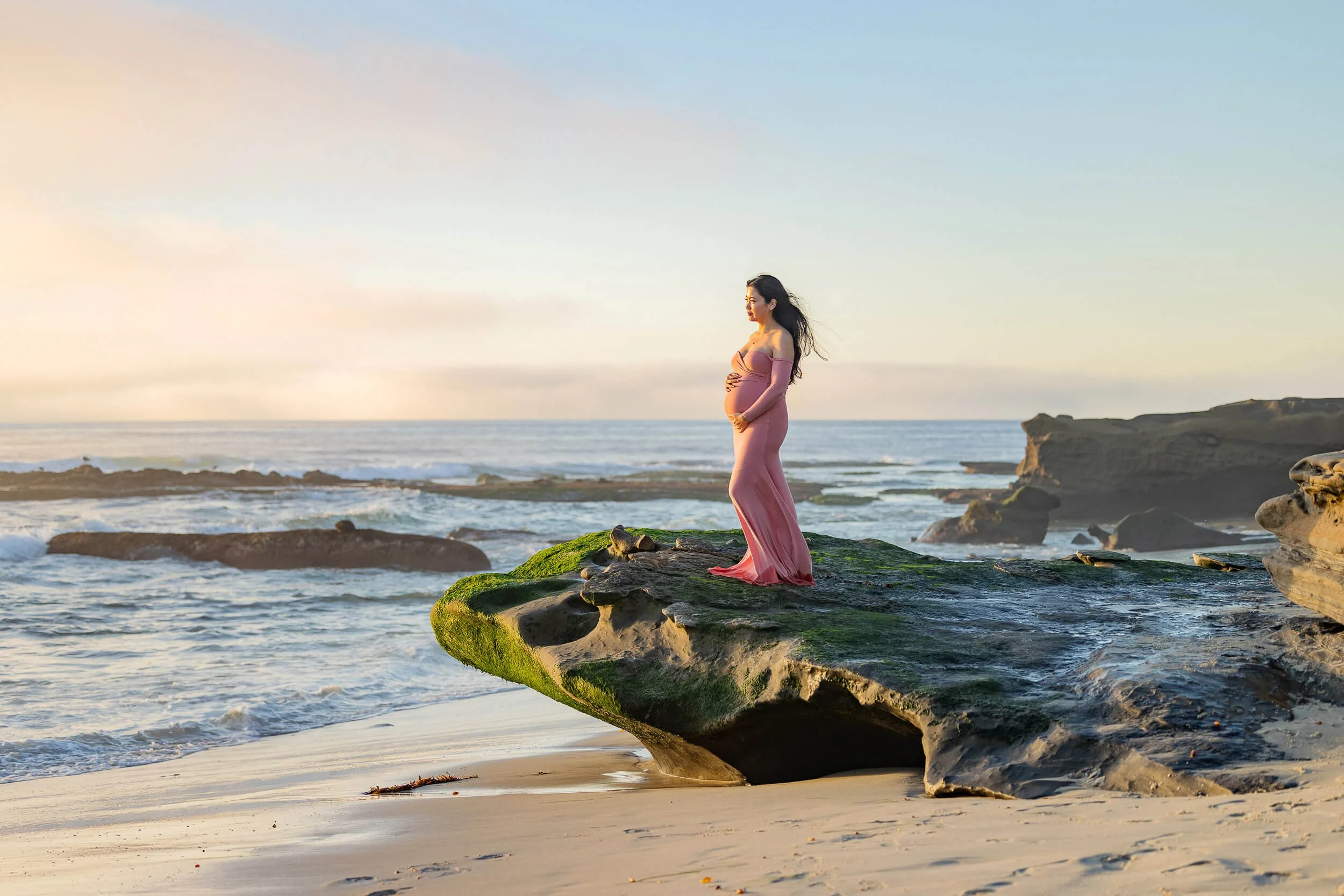 A pregnant woman in a pink dress standing on a moss-covered rock at the beach during sunset.