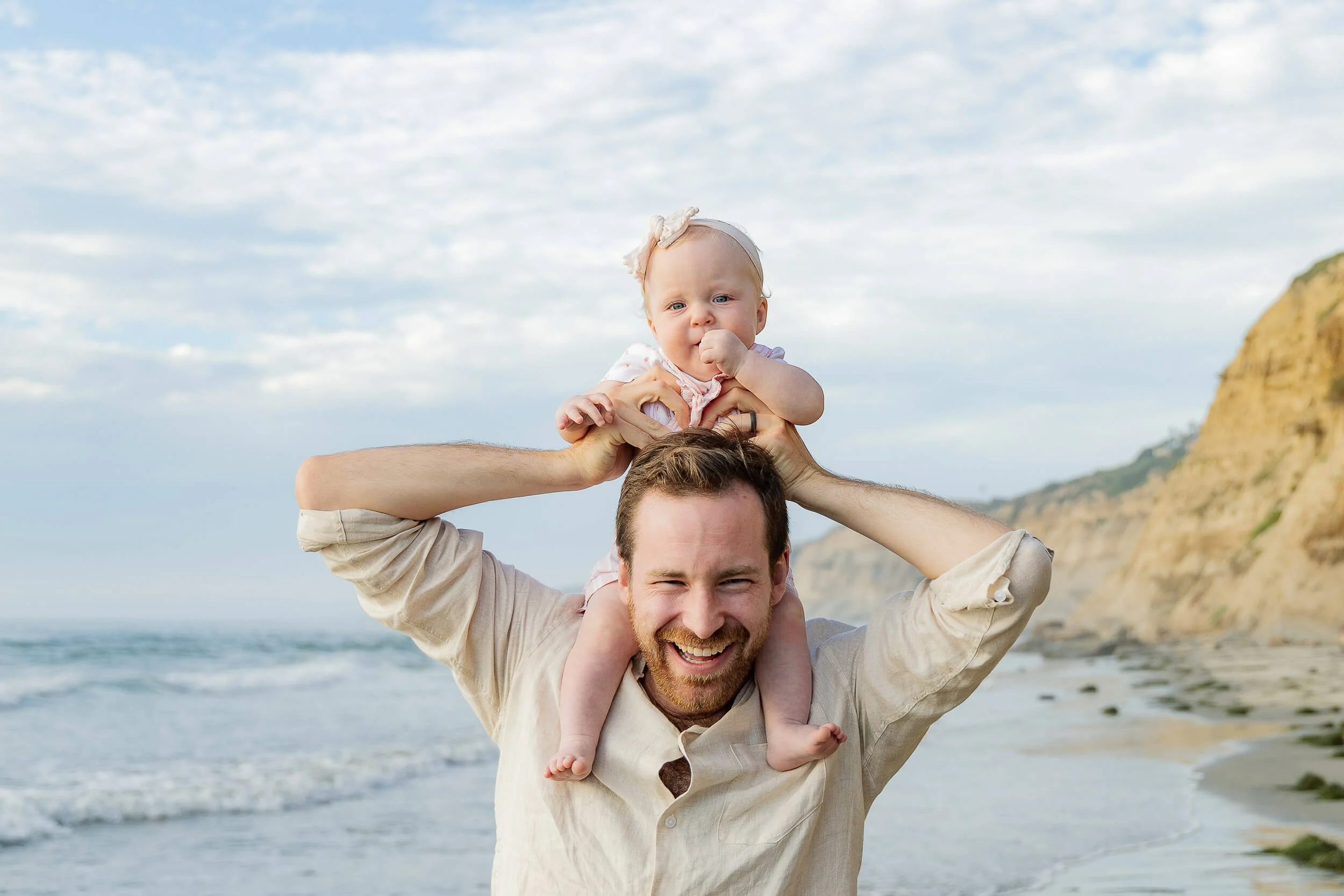 A man with a beard carrying a happy baby girl on his shoulders at the beach, with cliffs in the background and a partly cloudy sky.