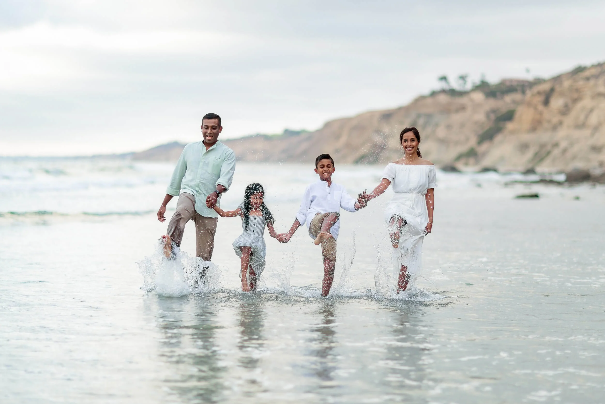 A family of five holding hands and running through the ocean water at the beach, smiling and splashing as they enjoy a day outdoors.