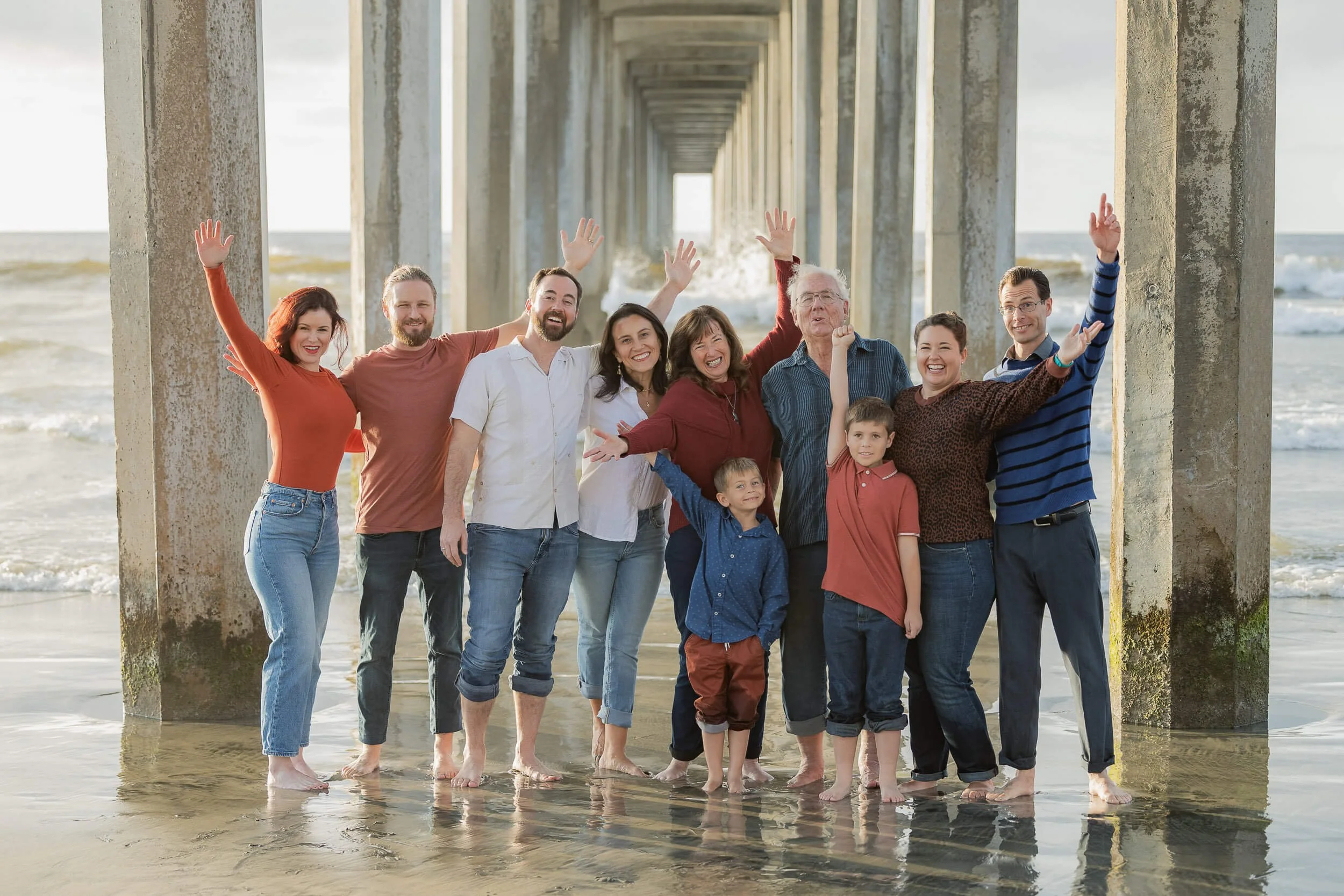 A large group of people, including children and adults, standing under a pier on a beach, smiling and raising their hands in celebration.