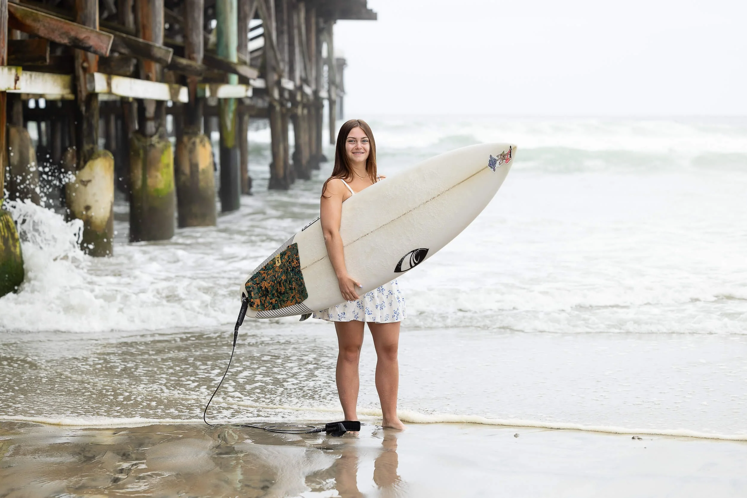 Woman in a white dress holding a surfboard at the beach near a pier, with ocean waves in the background.
