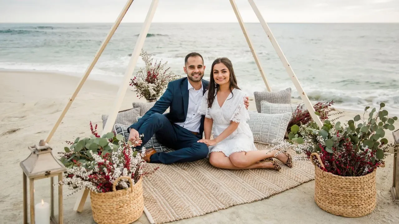 A couple sits on a woven mat on the beach under a wooden teepee, surrounded by flower arrangements and pillows, with the ocean in the background.