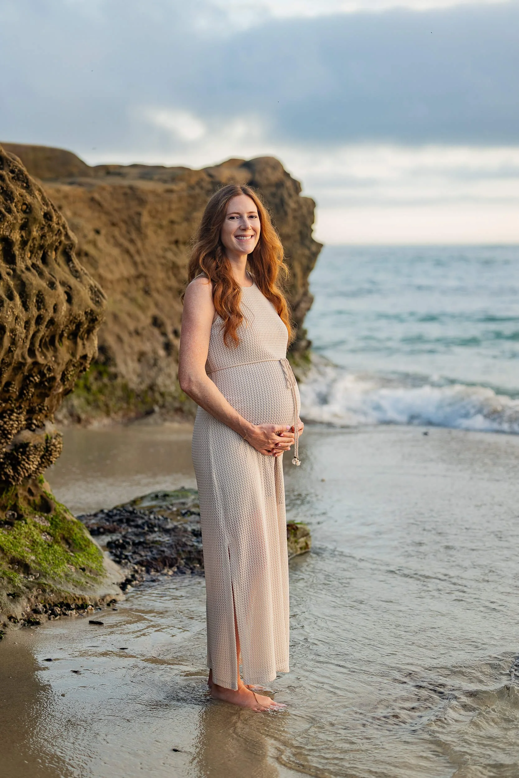 A pregnant woman standing on the beach near rock formations, wearing a sleeveless beige dress, smiling, with her hands resting on her belly.