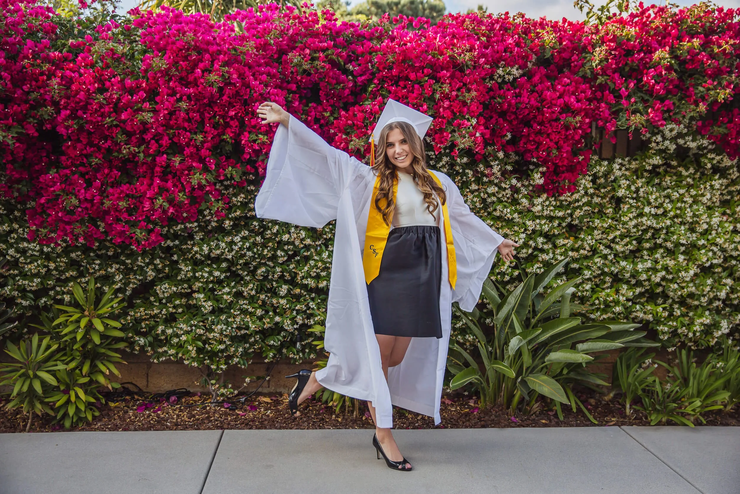 A young woman in a graduation cap and gown, with a yellow stole, posing with arms outstretched and smiling in front of a vibrant pink and white flowering bush.