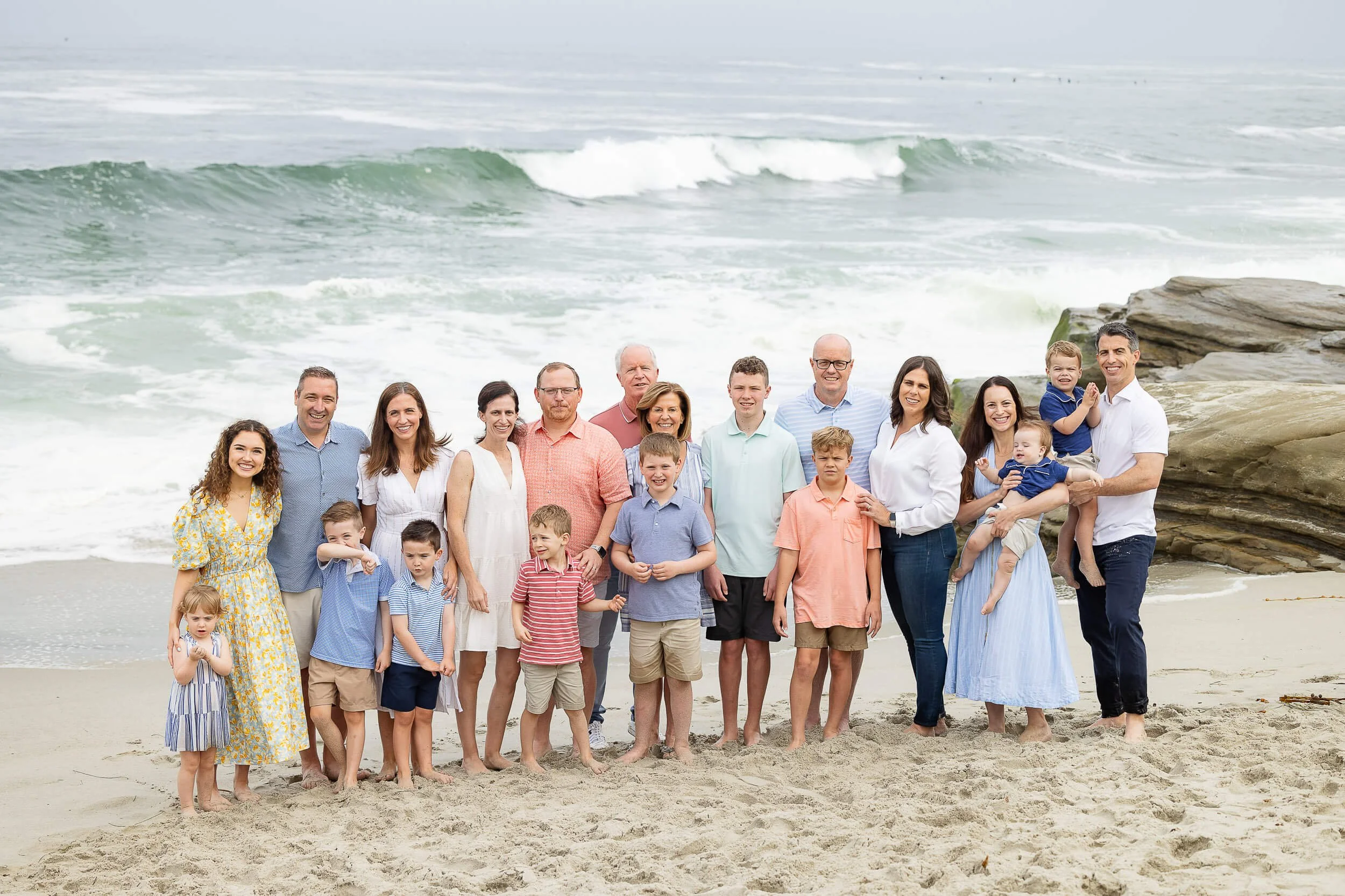 Family and friends gathered on a sandy beach near rocks with ocean waves in the background, posing for a group photo.