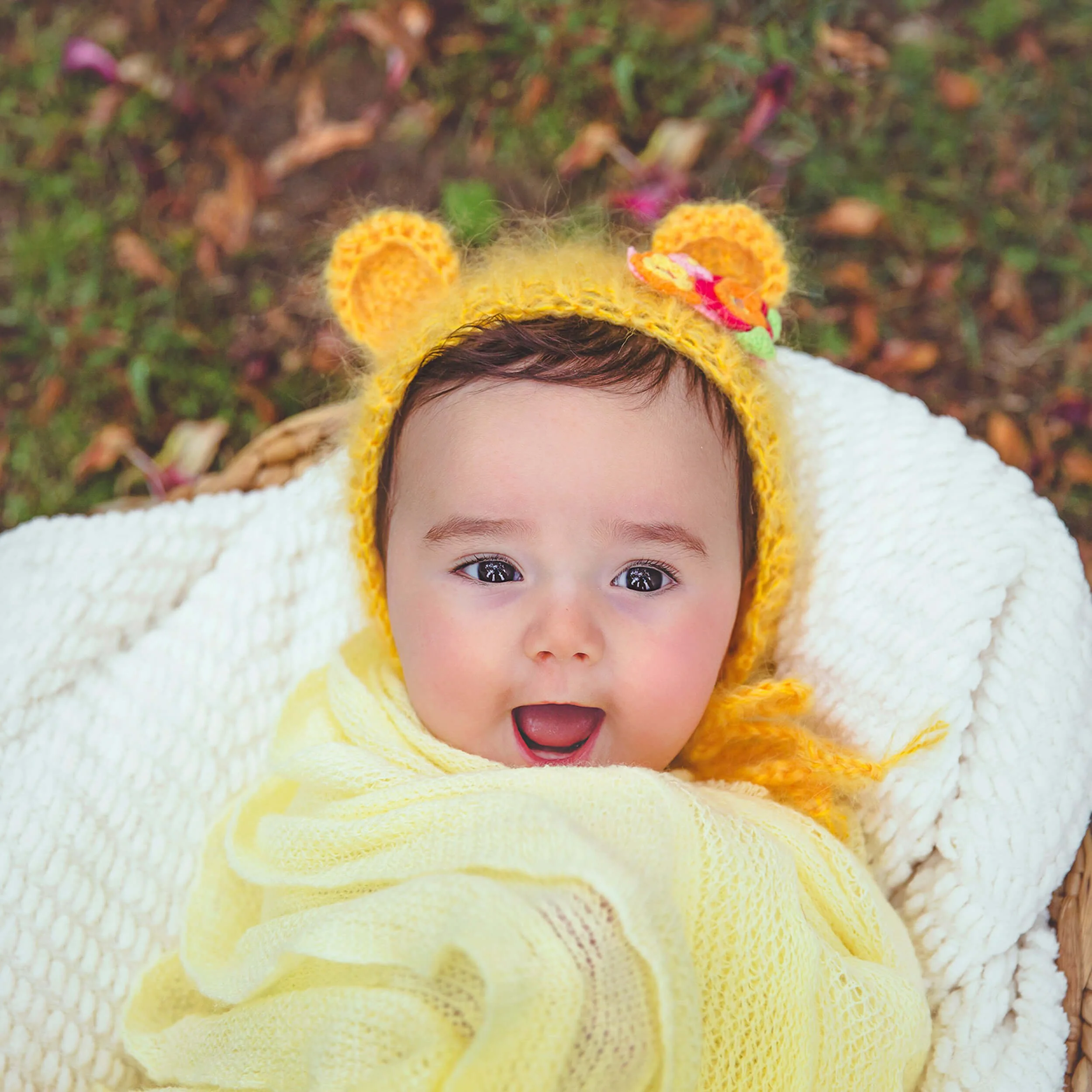 A baby with dark hair, wearing a yellow bear hat and a yellow blanket, lying on a white textured blanket outdoors, surrounded by fallen leaves.