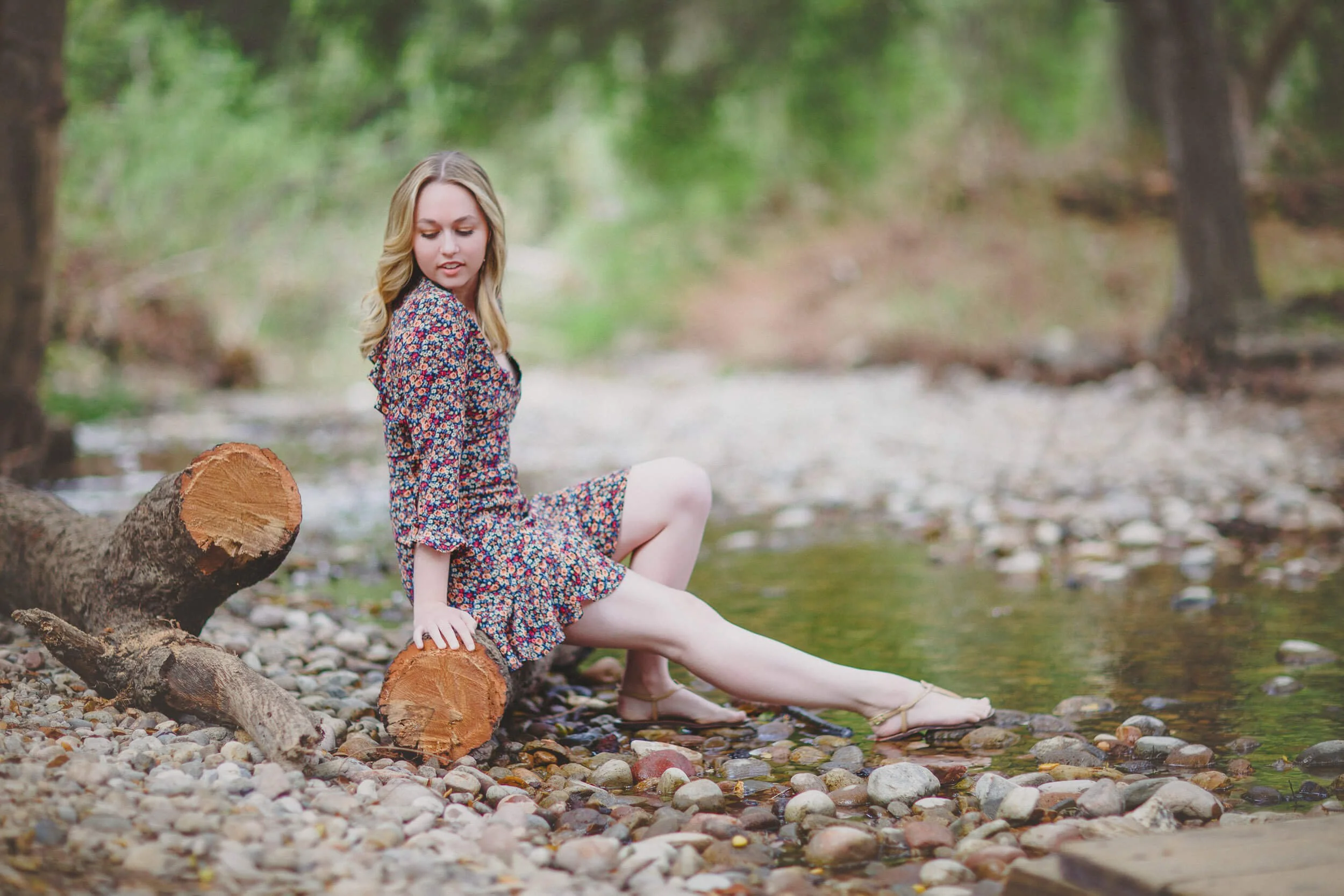 A young woman in a floral dress sitting on a log by a creek in a forested area.
