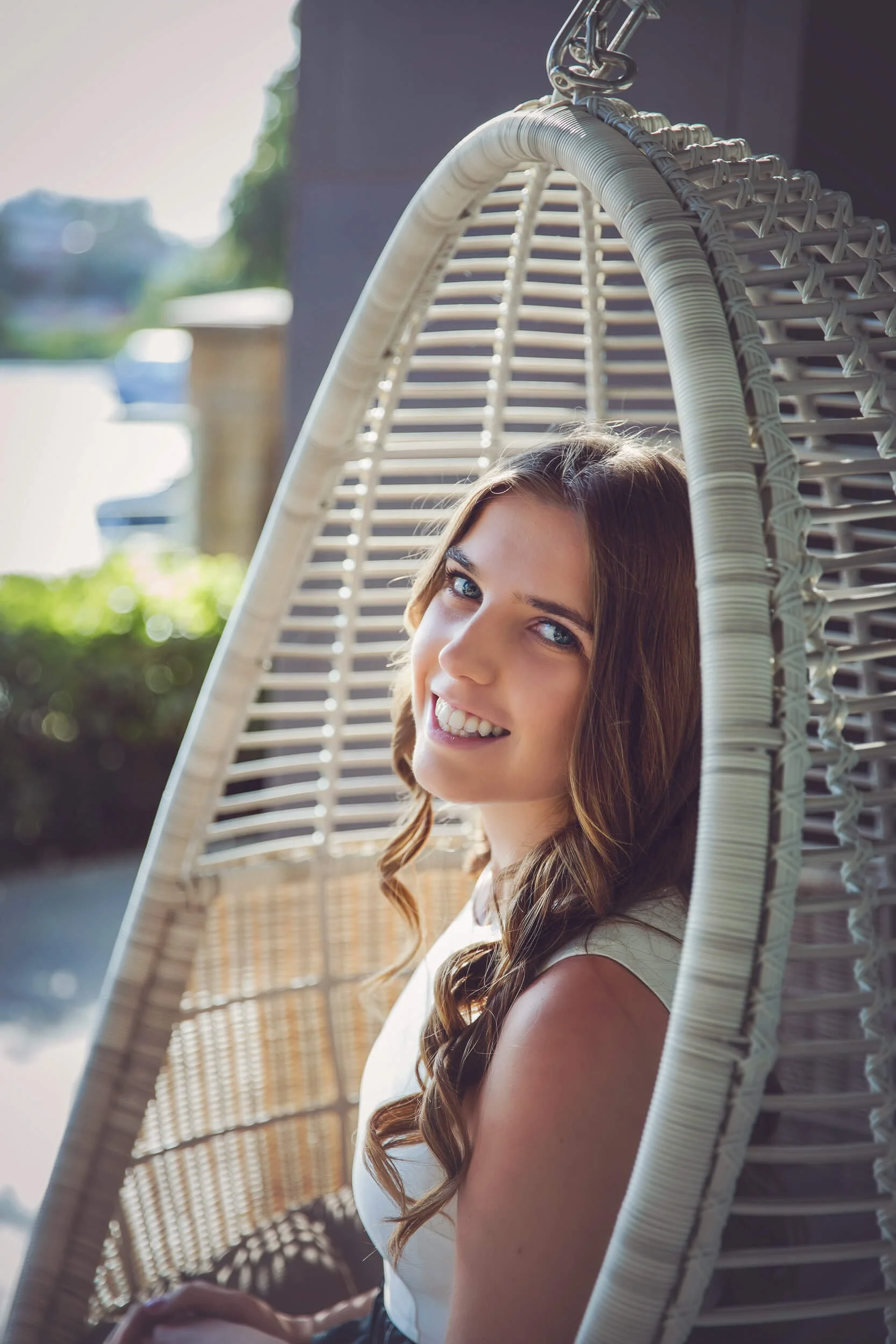 A young woman with long brown hair sitting in a hanging wicker chair, smiling at the camera. She is outdoors with a blurred background of greenery and buildings.