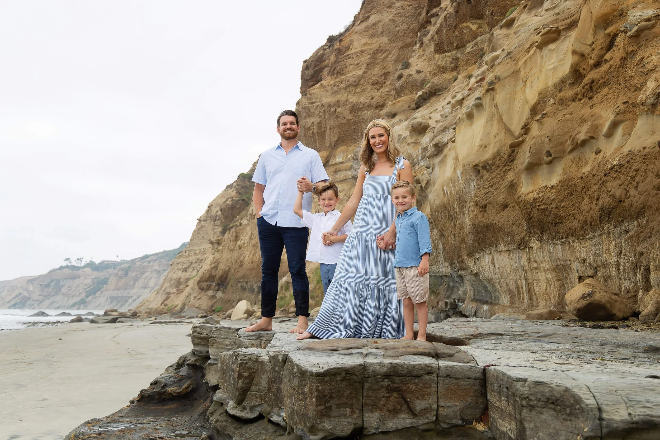 A family of four standing on a rock at the beach near cliffs, smiling at the camera. The mother and children are holding hands, with the father standing nearby. The sky is overcast.