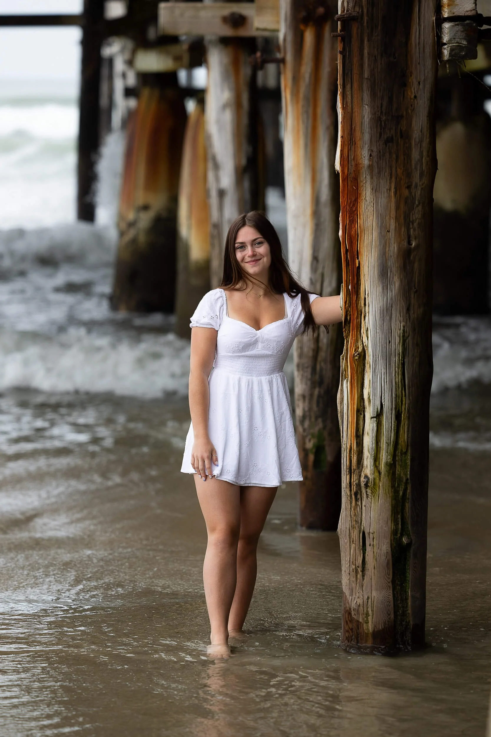 A woman in a white dress standing in shallow water under a wooden pier, smiling at the camera.