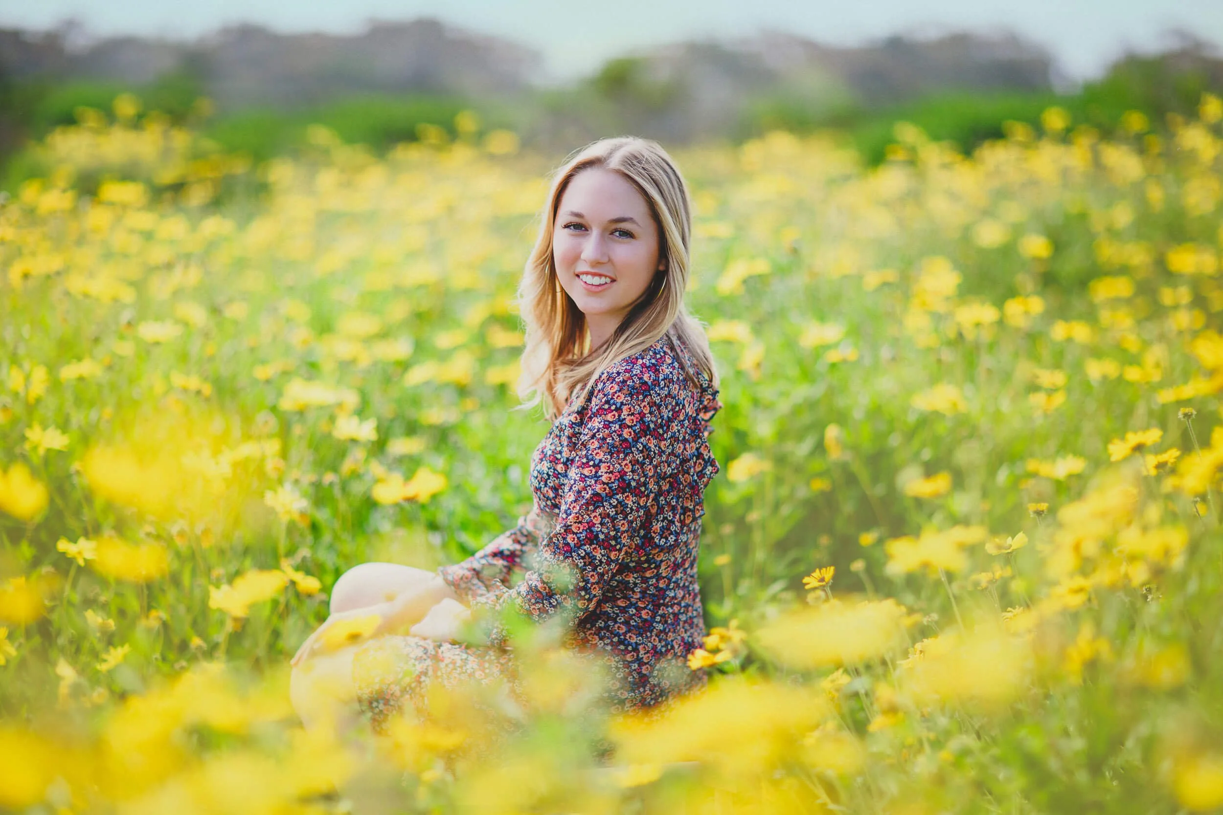 A young woman with blonde hair smiling while sitting in a field of yellow flowers.