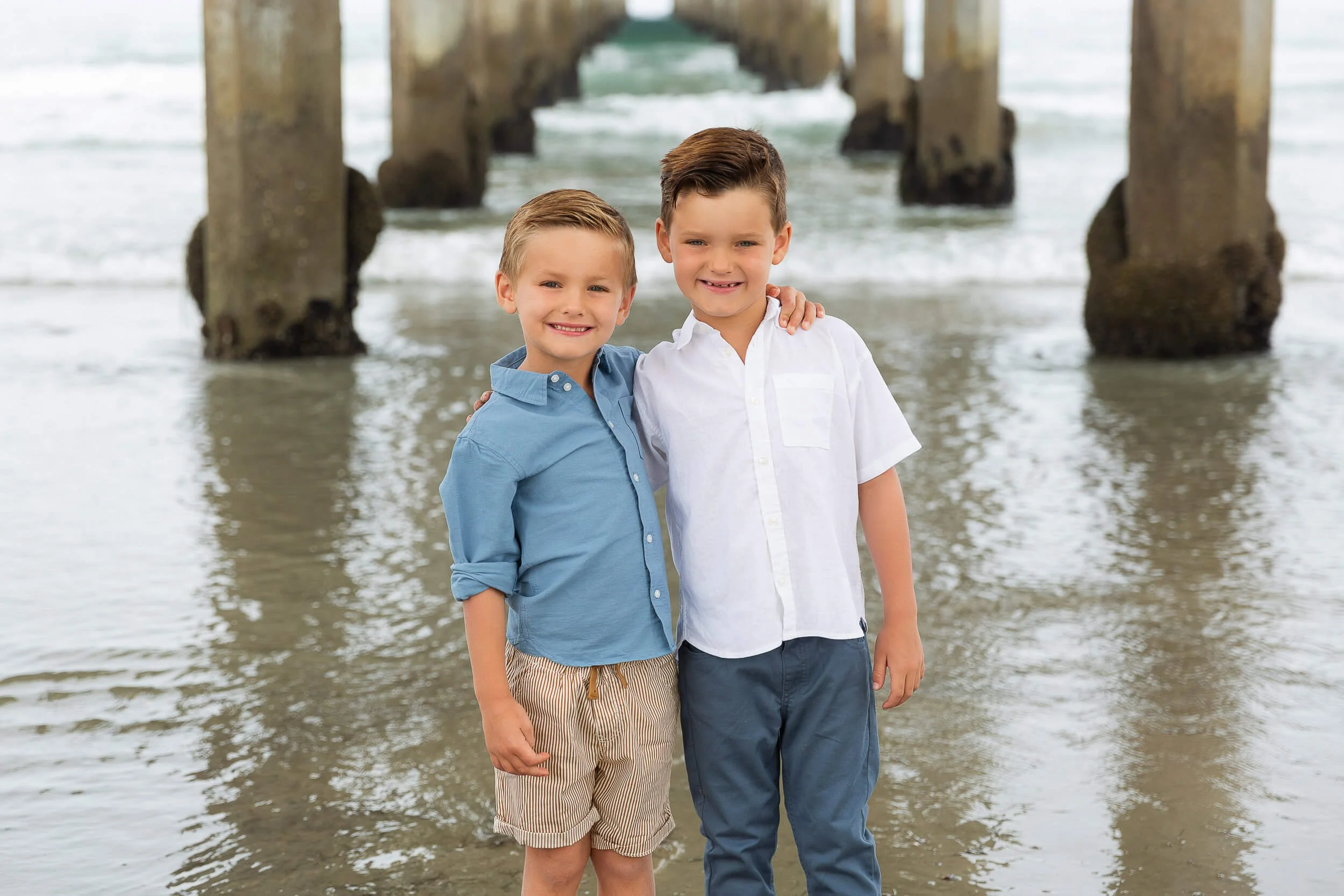 Two young boys standing close together on a beach under a pier, smiling at the camera, with water and pier pillars in the background.