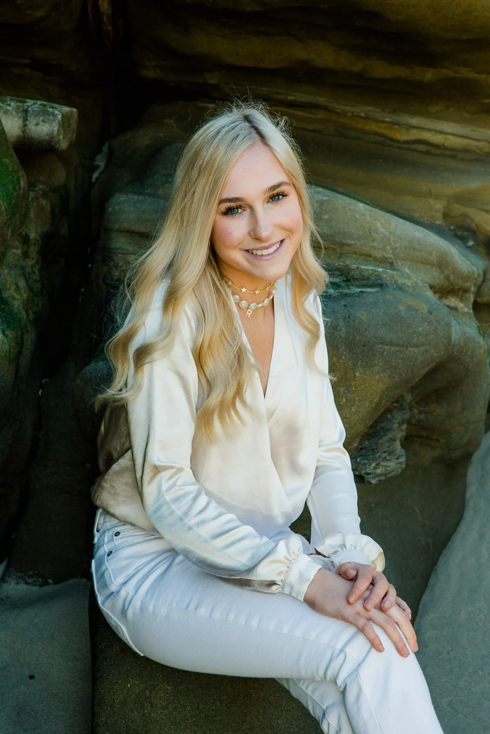 A young woman with long blonde wavy hair, wearing a cream satin blouse and white pants, sitting on rocks at the beach and smiling at the camera.
