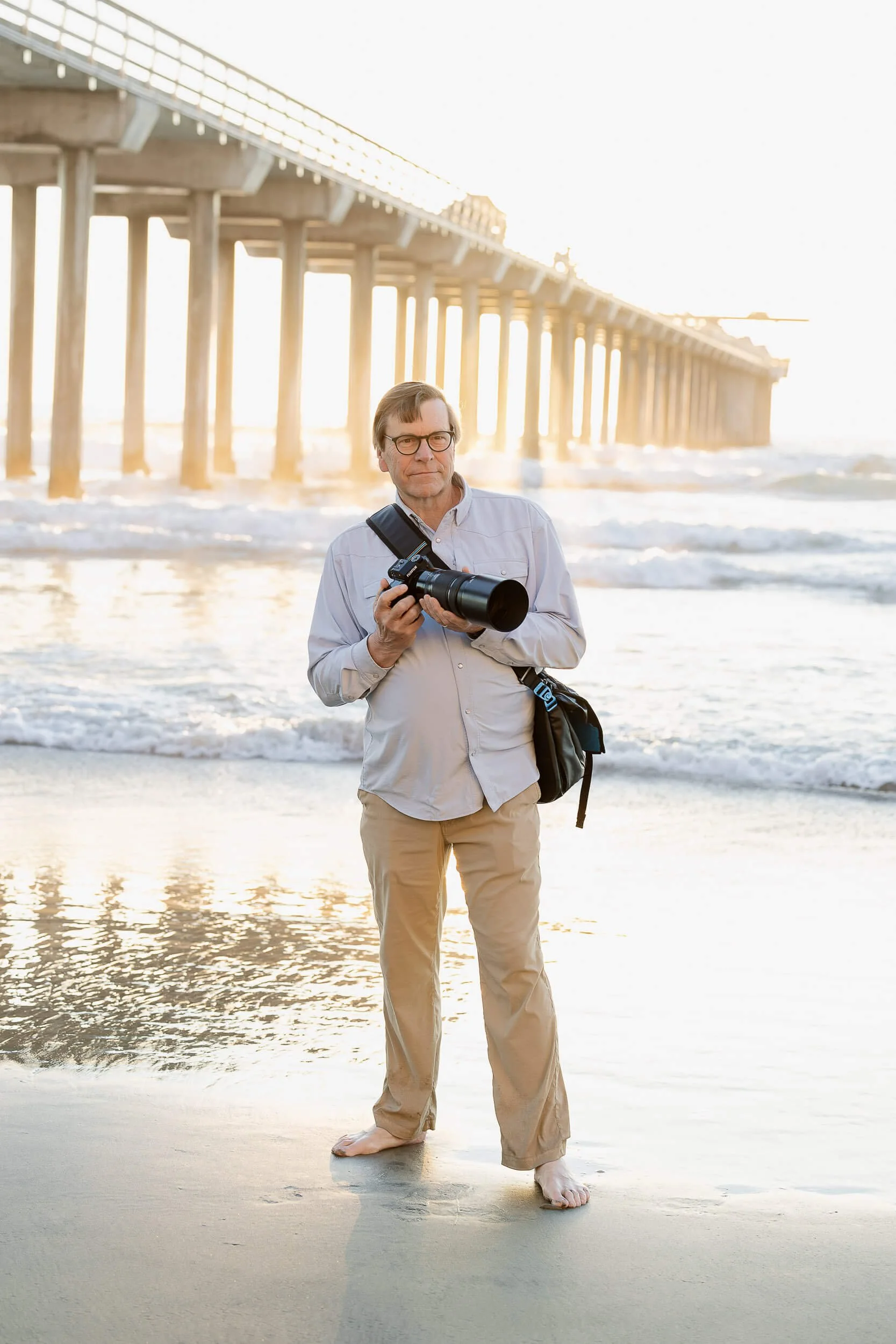 A man standing on the beach with a camera, near a pier, during sunset.