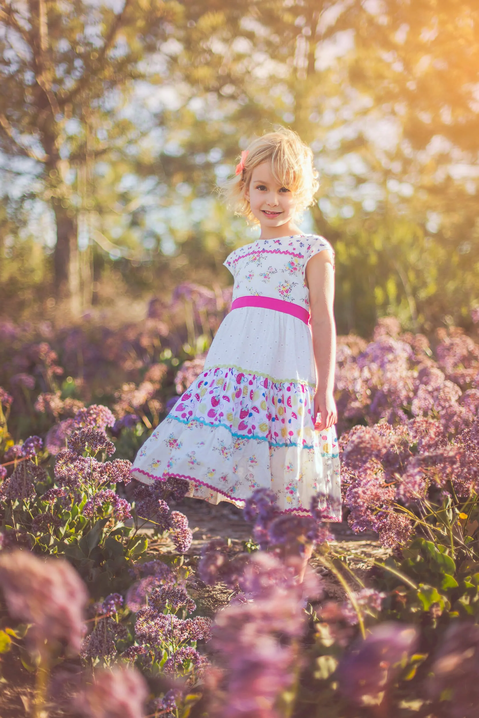 A young girl in a white, colorful dress standing among purple flowers in a garden during golden hour sunlight.