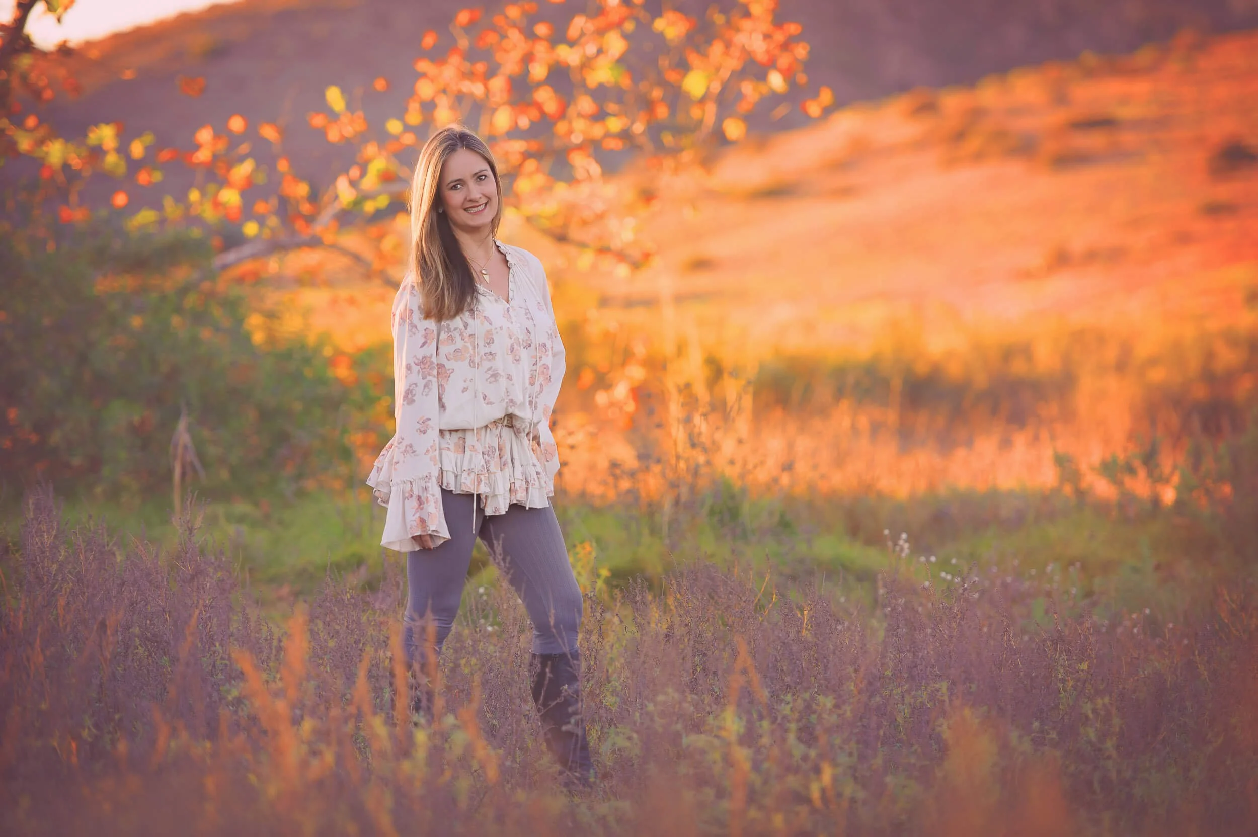A smiling young woman with long brown hair stands in a field of purple flowers during autumn, with orange trees and a sunset sky in the background.