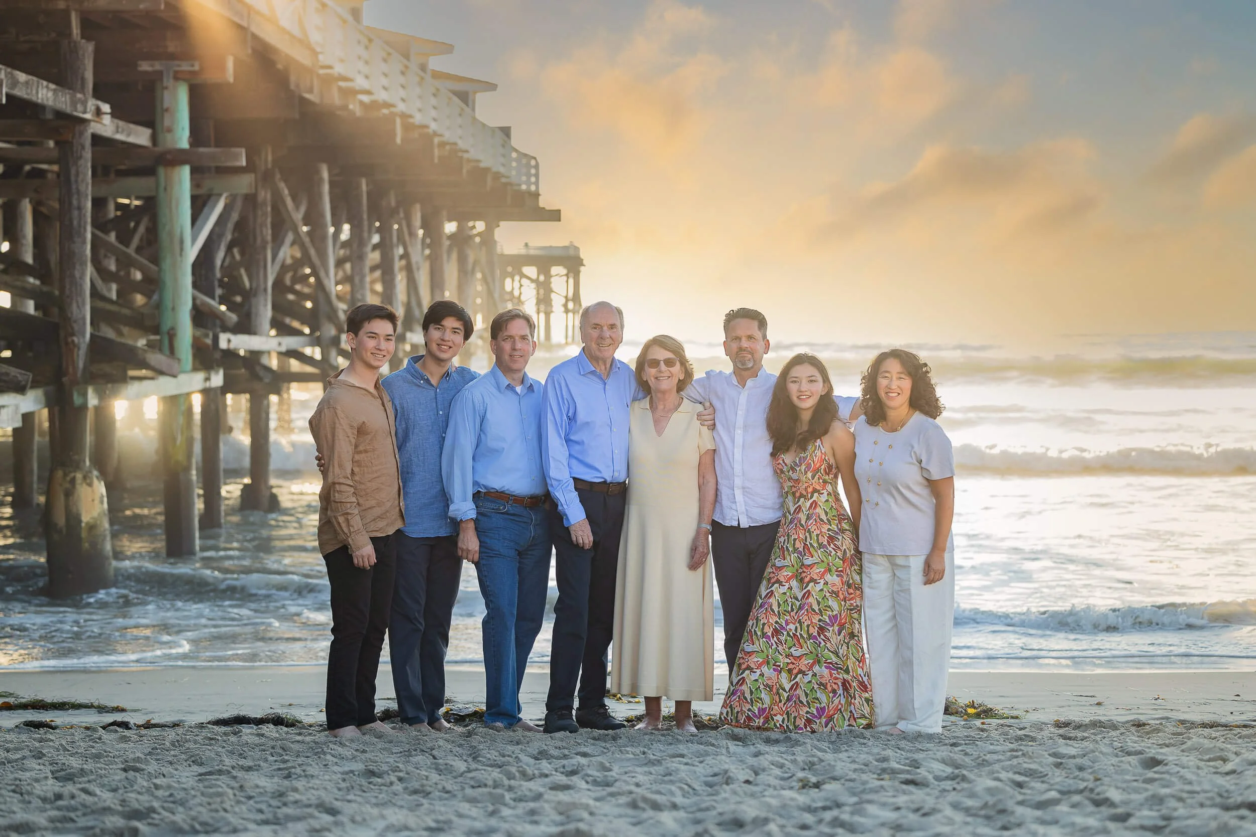 A family of nine posing on a beach at sunset in front of a pier with the ocean waves in the background.