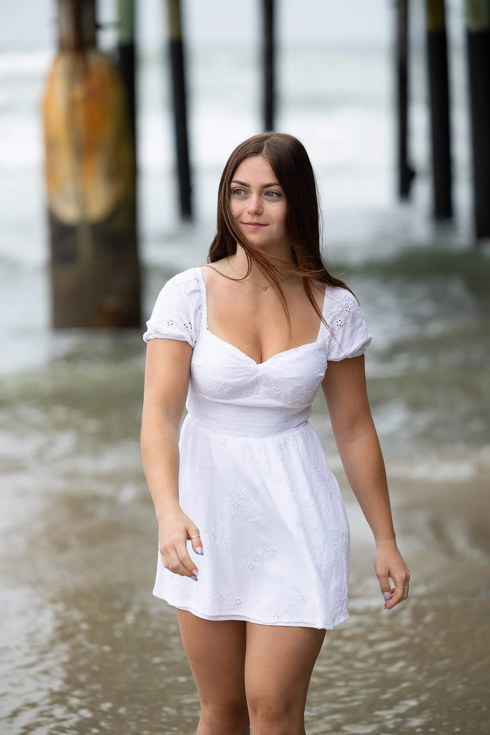 A young woman in a white dress standing in the water near a beach with a pier in the background.