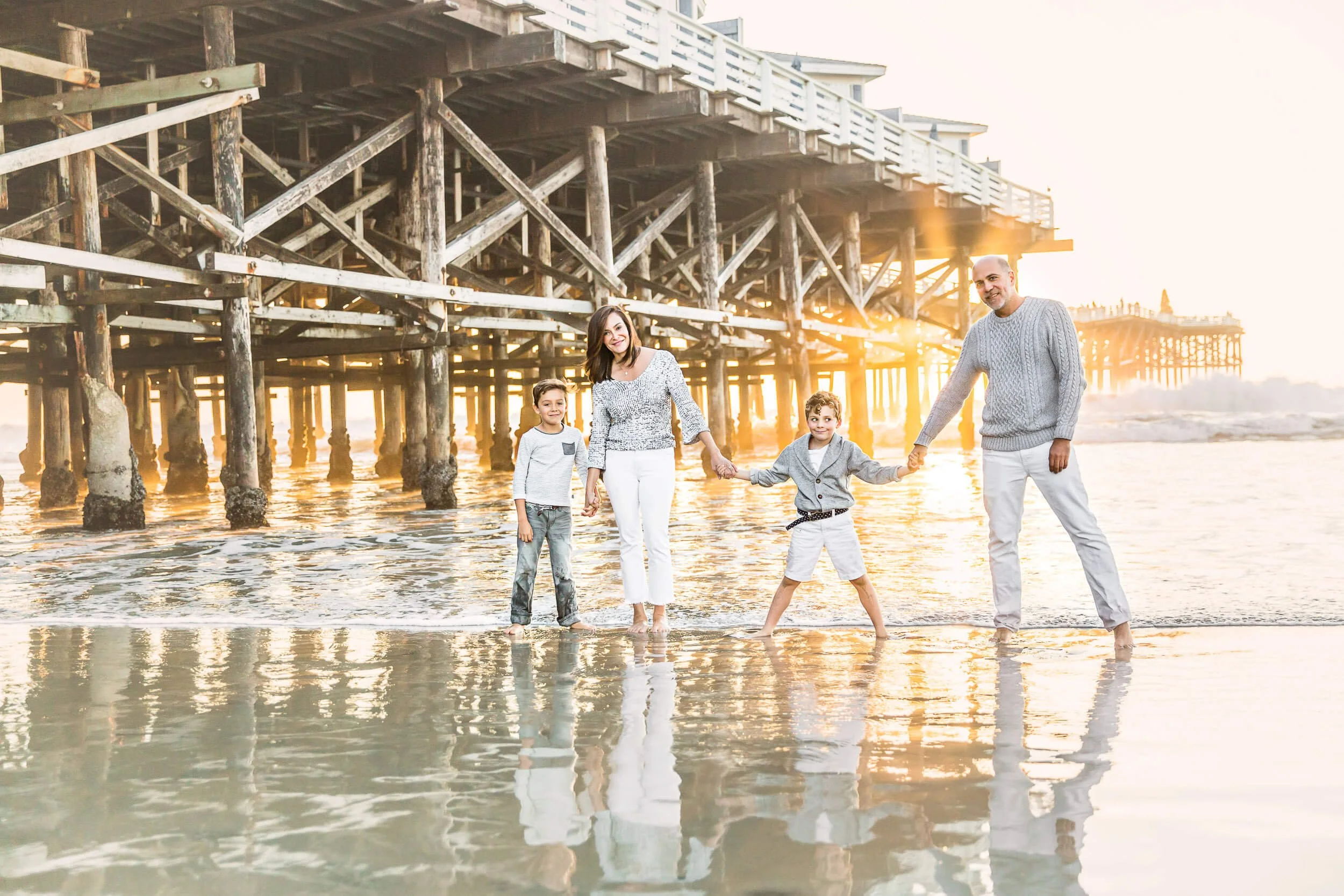 A family of four holding hands on a beach at sunset, with a wooden pier in the background.