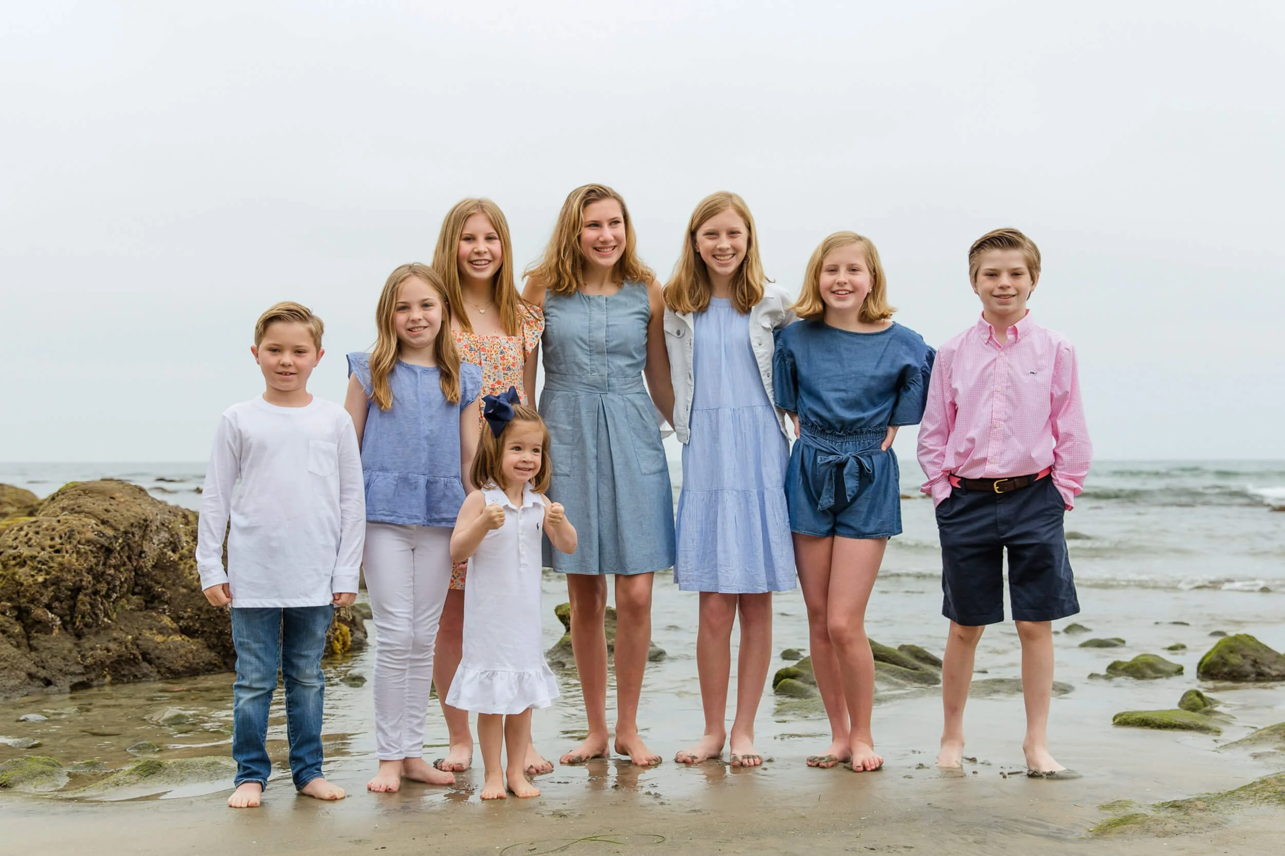 A group of ten children, mostly girls and one boy, standing barefoot on the beach in a line near the rocks with the ocean in the background, smiling.