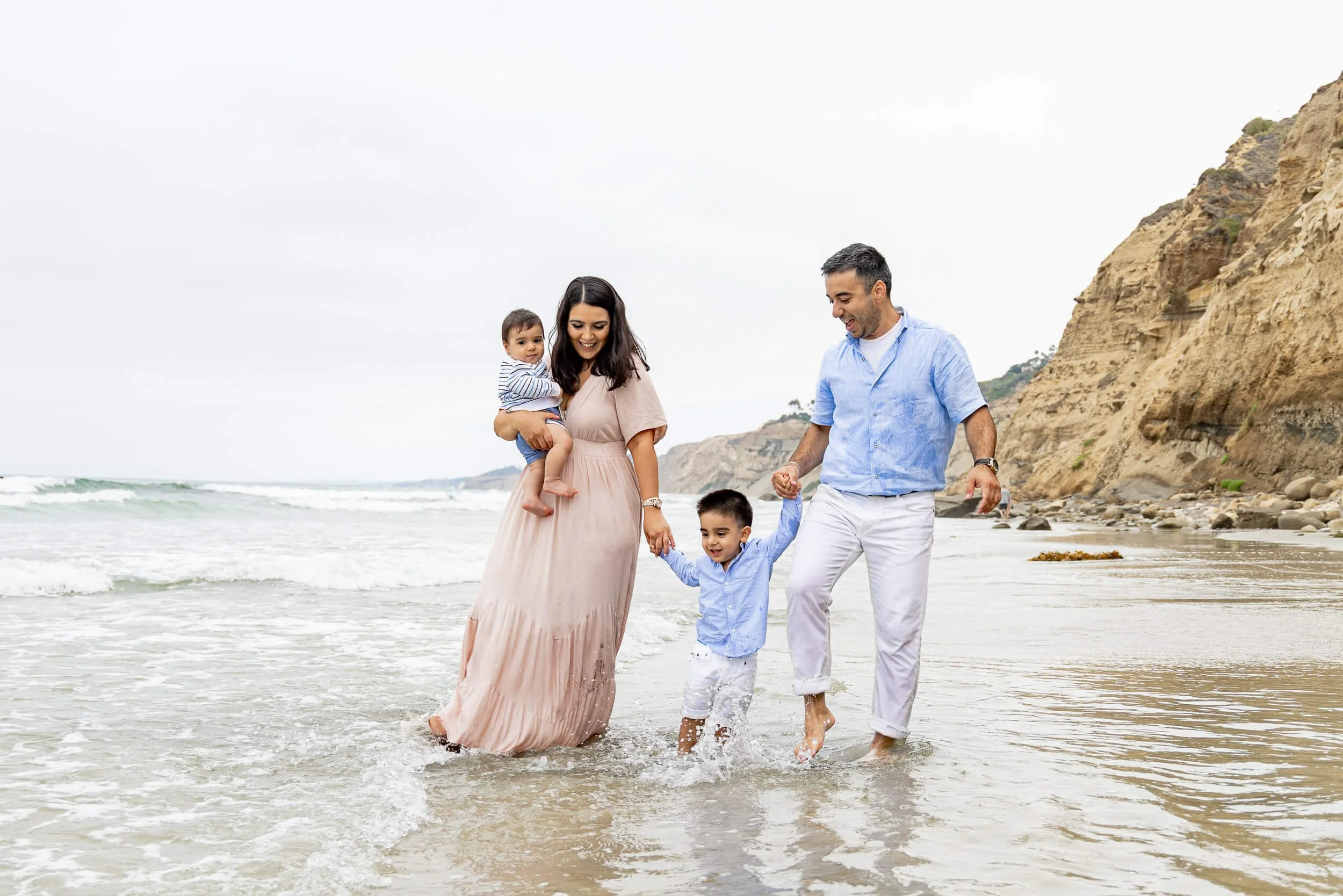 A family of four enjoying a walk on the beach, holding hands and splashing in the shallow water, with cliffs in the background.