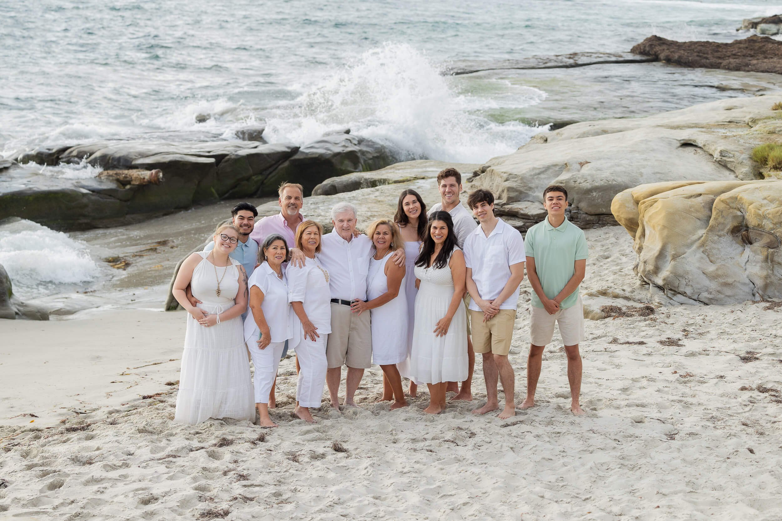 Group of people on a beach, standing on sand, with rocks and waves in the background, smiling for a photo.