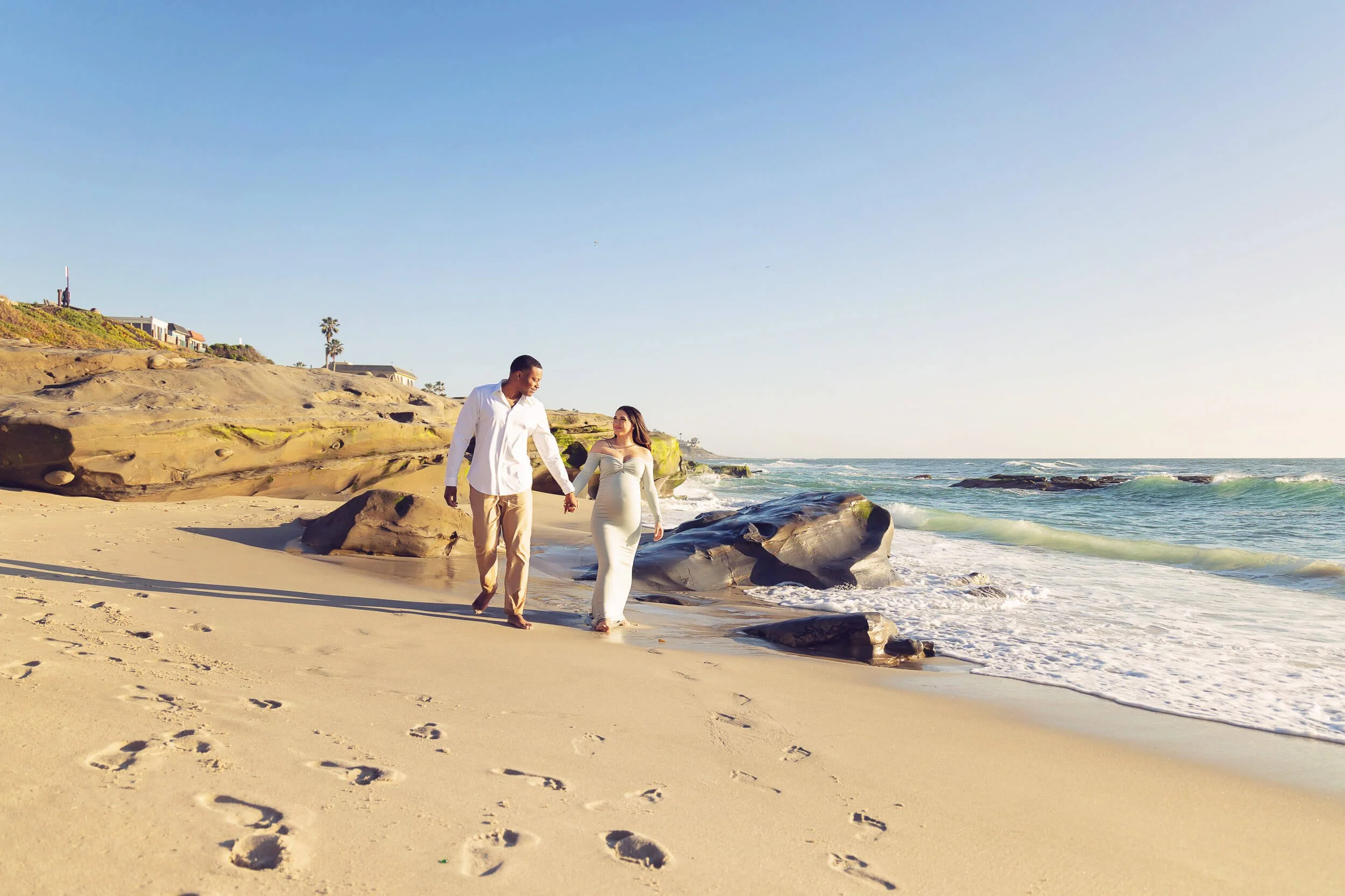 A couple holding hands walking on the sandy beach near the ocean with rocks and cliffs in the background.
