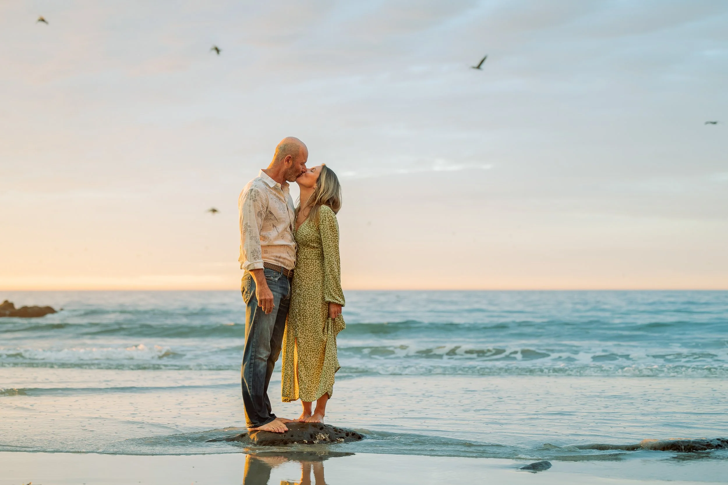 A couple kissing on a rock in the ocean at sunset, with seagulls flying overhead.
