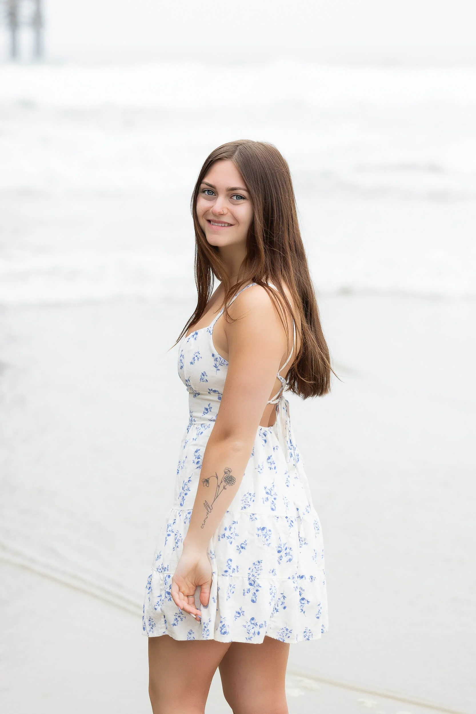 A young woman with long brown hair, wearing a white dress with blue floral patterns, standing on a beach with the ocean in the background, smiling at the camera.