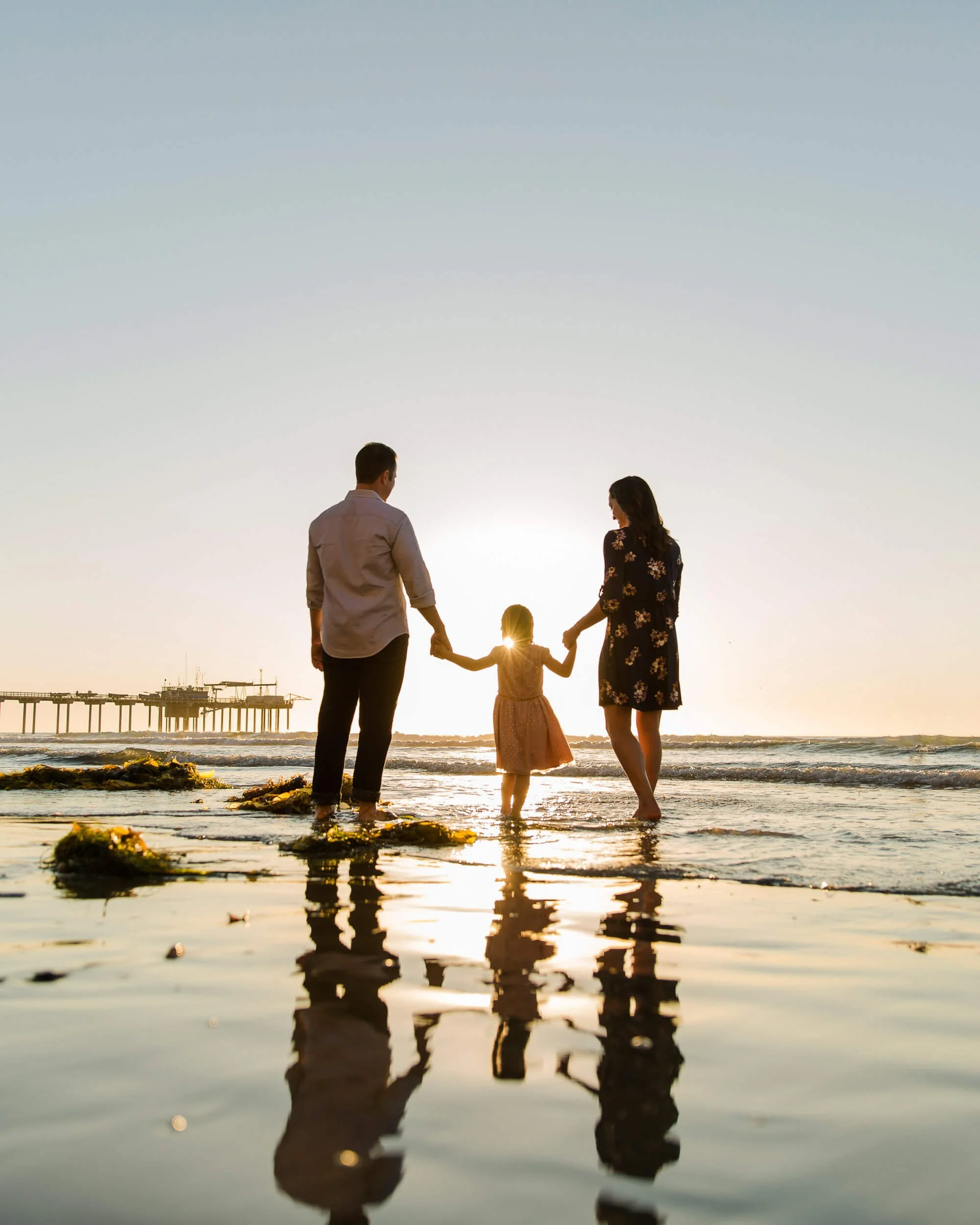 A family of three holding hands at the beach during sunset, with a pier in the background and their reflections in the water.