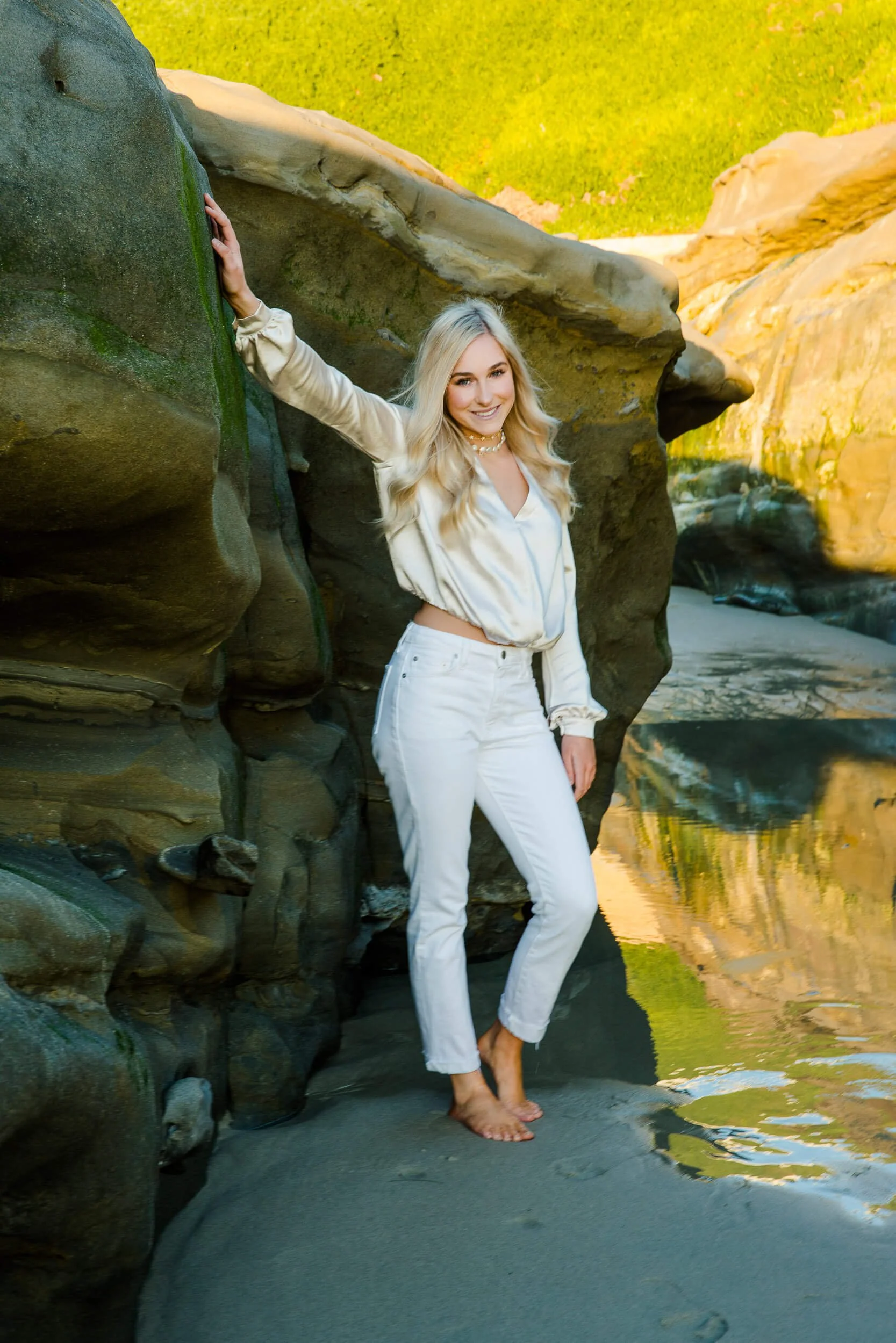 A woman with long blonde hair wearing a silky beige shirt and white jeans, standing barefoot on a sandy beach next to rocks, smiling at the camera.