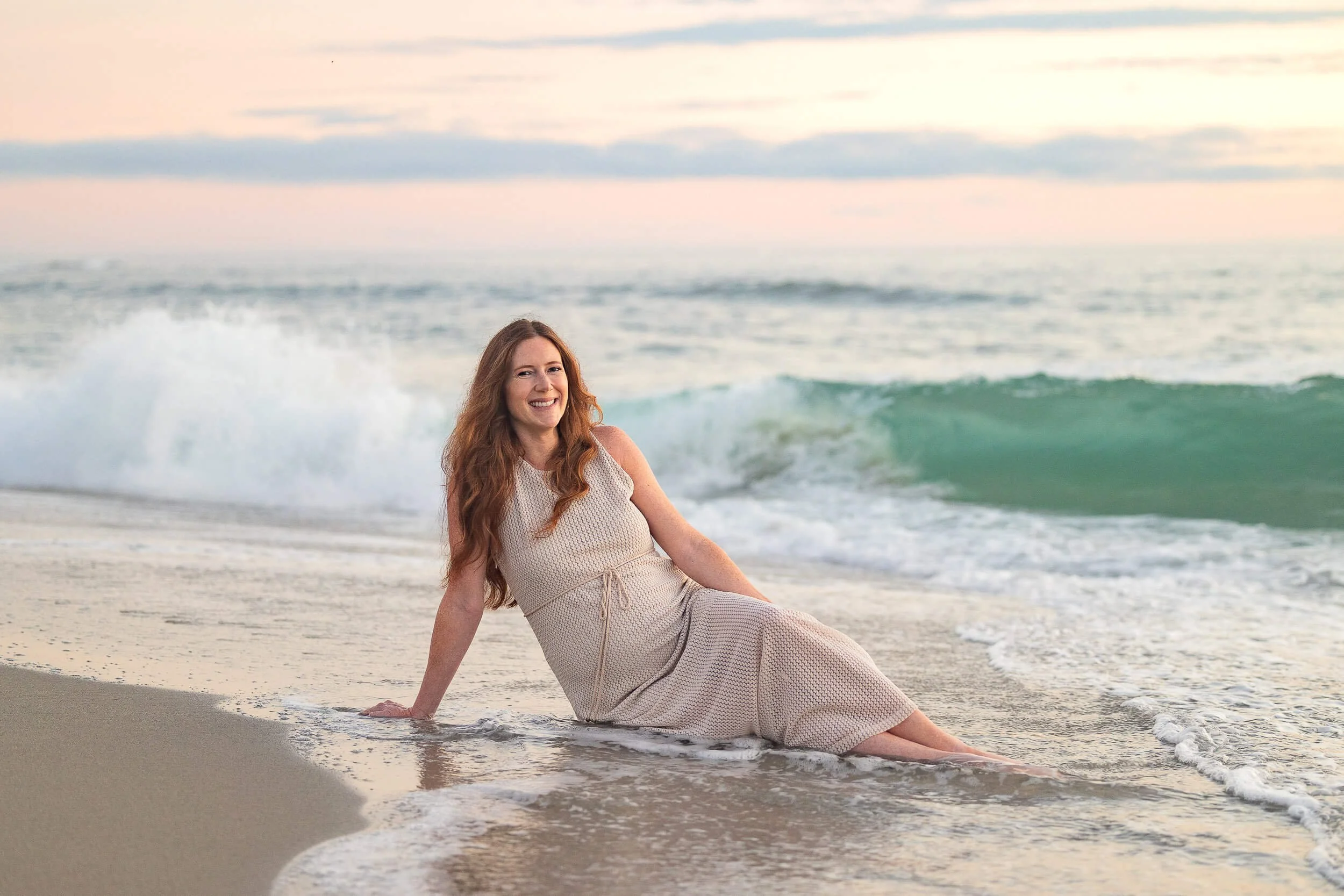A woman sitting in shallow ocean water on a beach during sunset, smiling and looking at the camera, with waves and a cloudy sky in the background.