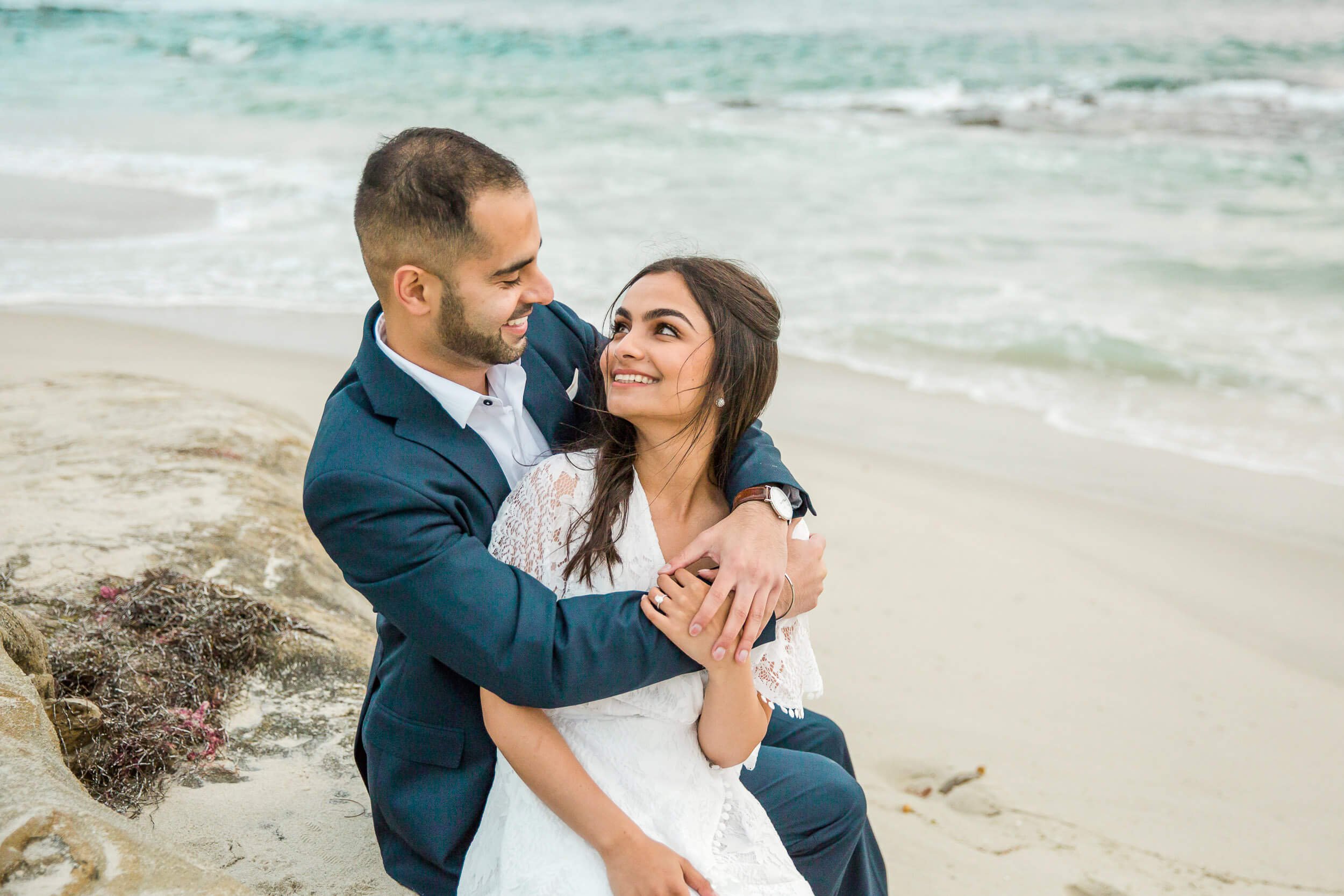 A couple in formal attire sitting on the beach, embracing and smiling at each other with the ocean in the background.
