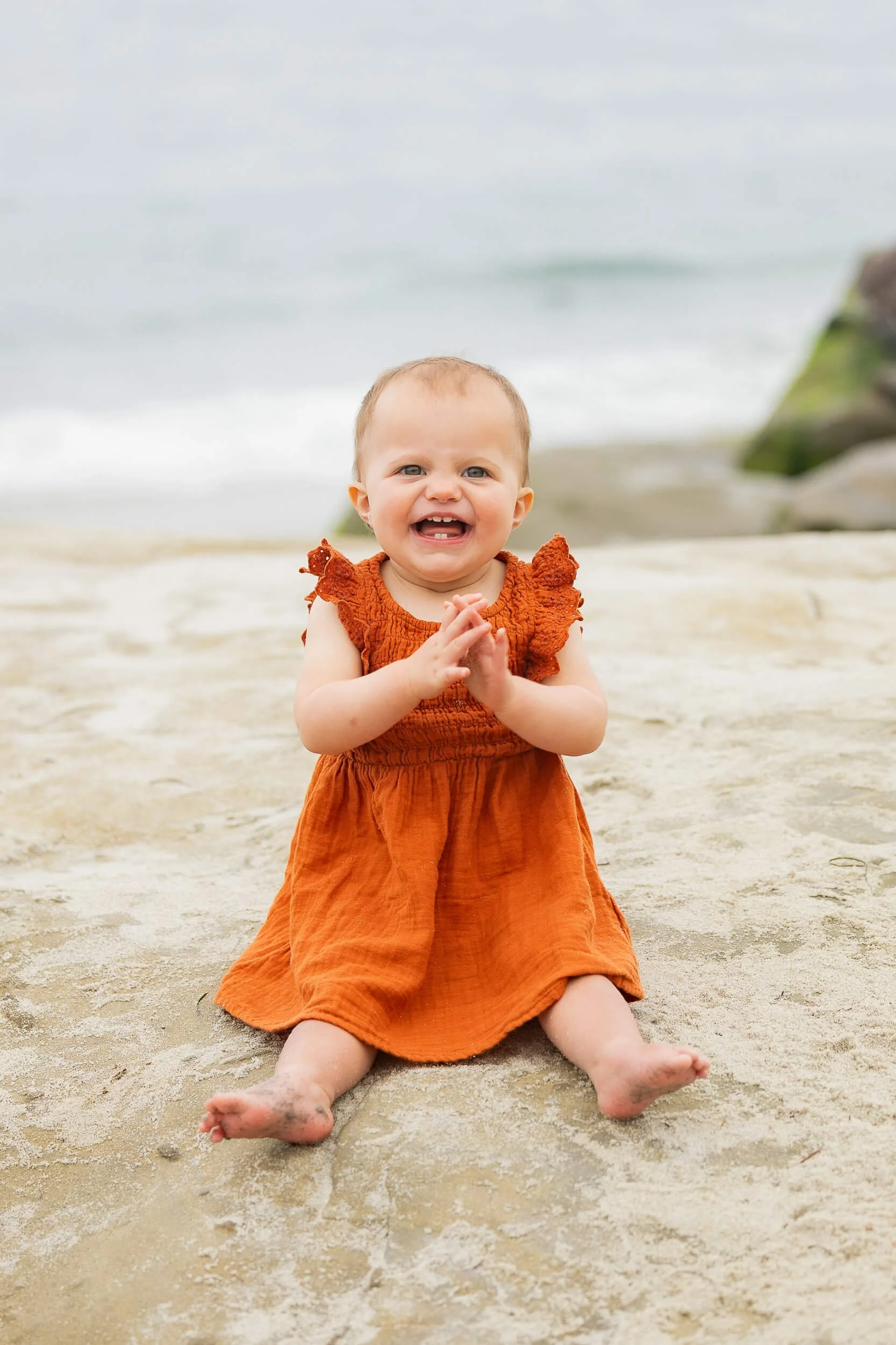 A young child sitting on a sandy beach, smiling with hands clasped, wearing an orange dress with ruffled sleeves, with the ocean and rocks in the background.