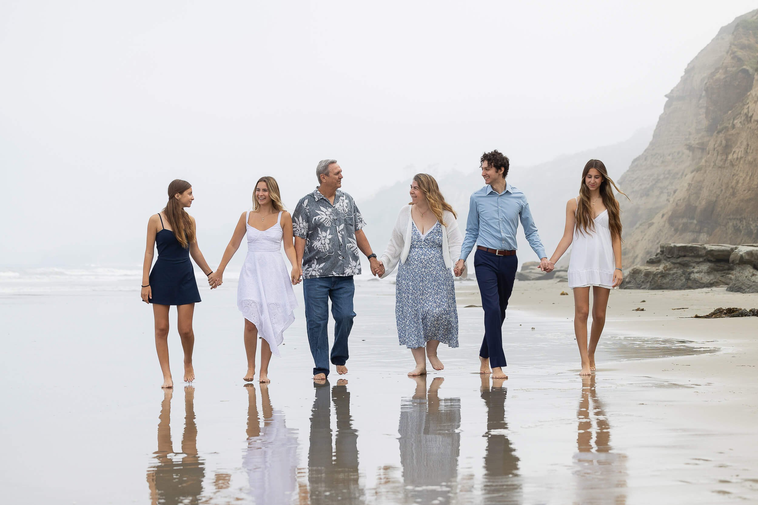A family of seven holding hands walking along the beach in foggy weather, with cliffs in the background.