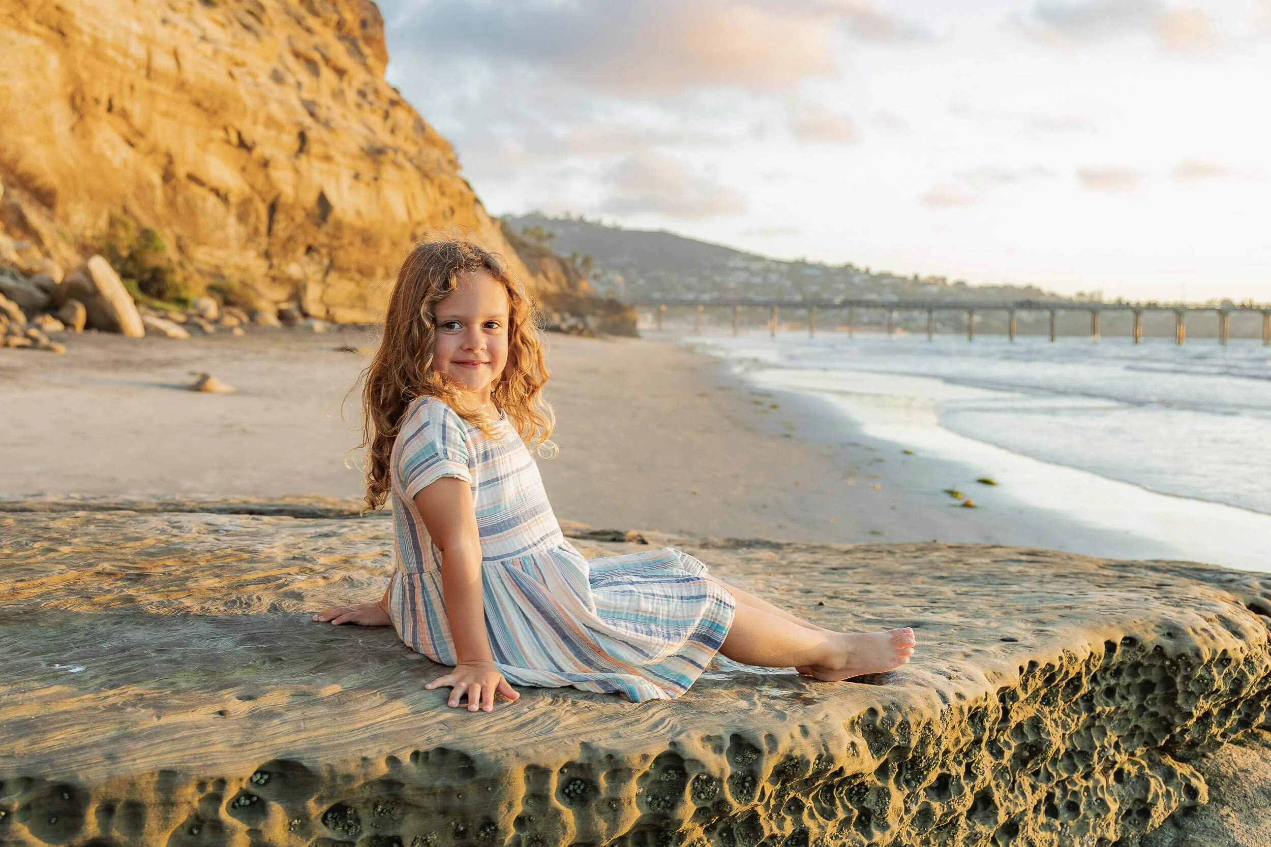 A young girl with curly hair in a striped dress sitting on a large rock on the beach during sunset.
