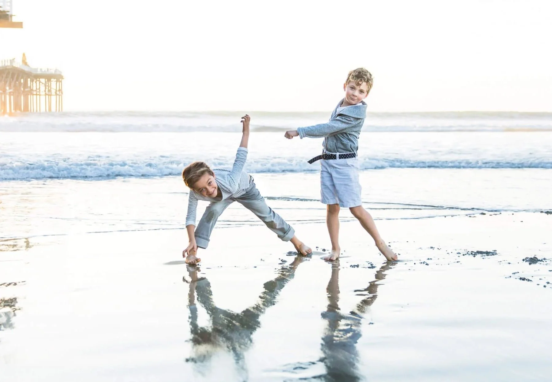 Two young boys playing on the beach with ocean waves in the background, one bending down and smiling, the other standing and pointing at him, both dressed in casual clothes.