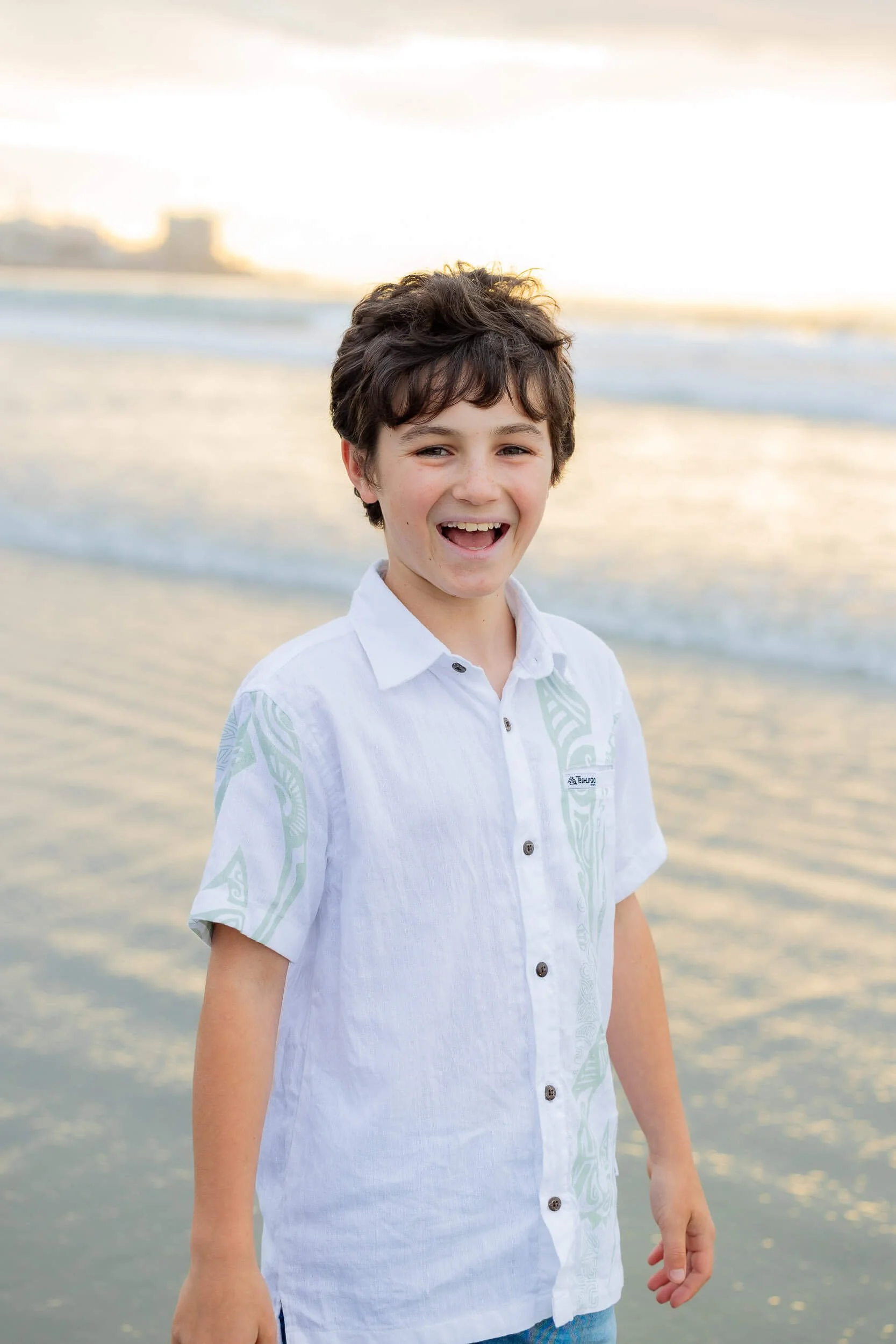 A young boy smiling and laughing at the beach during sunset, standing in shallow water with waves in the background.