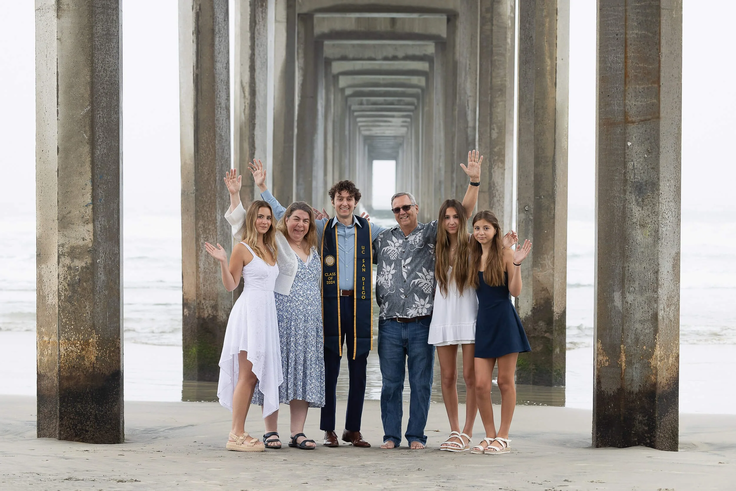 A family standing under a pier on the beach, celebrating a graduation with one person wearing a graduation gown and cap.