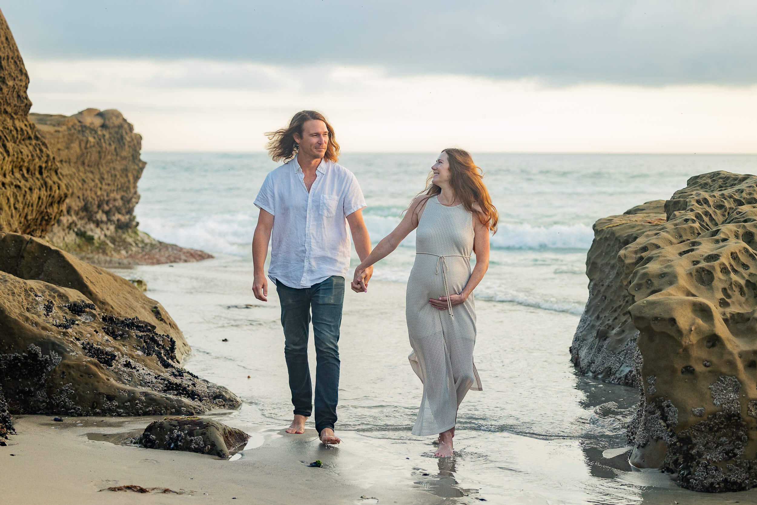 A couple walks hand in hand along the beach near rocks, with the ocean waves in the background, under an overcast sky.