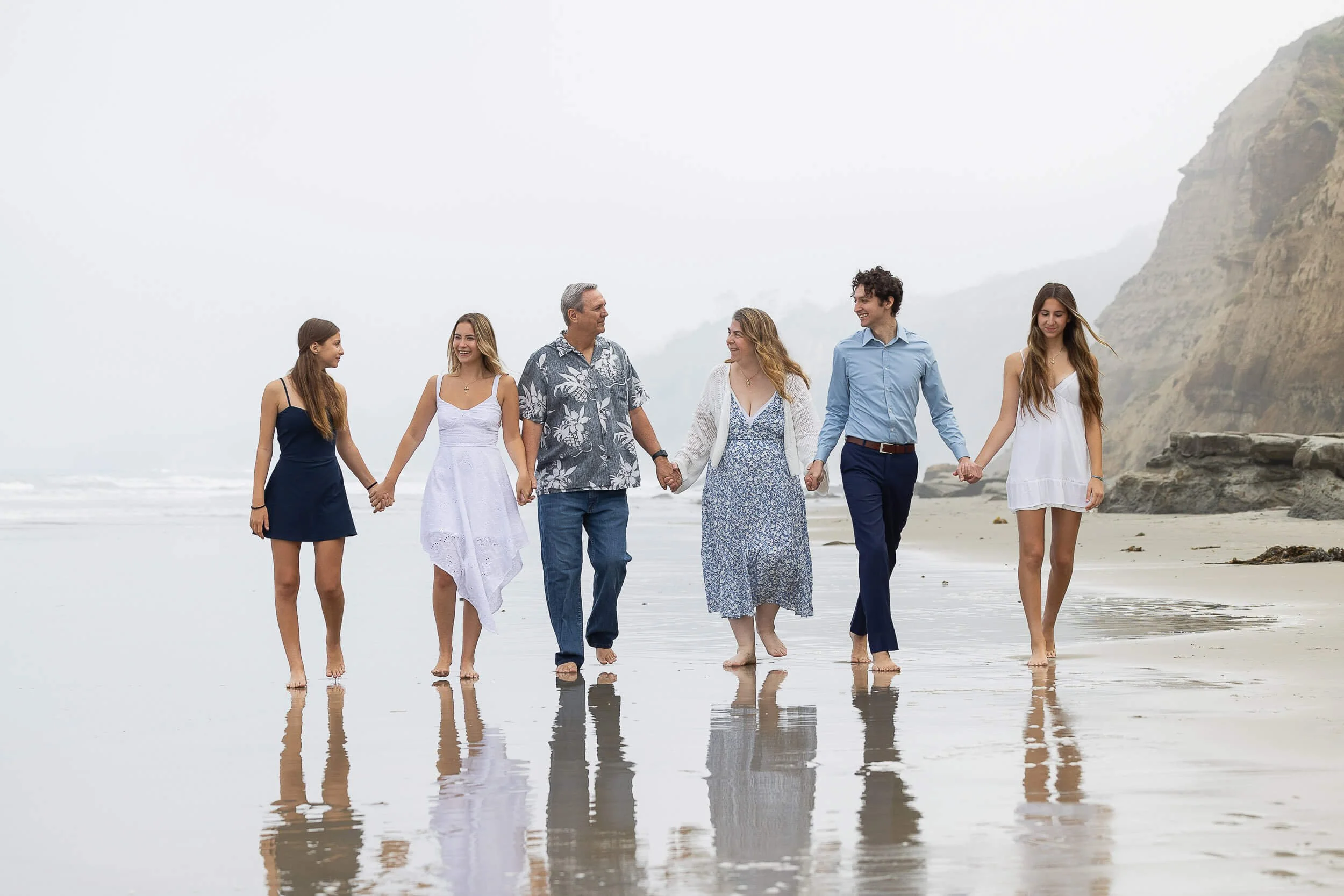 Family holding hands and walking on the beach in the foggy seaside with rocky cliffs.
