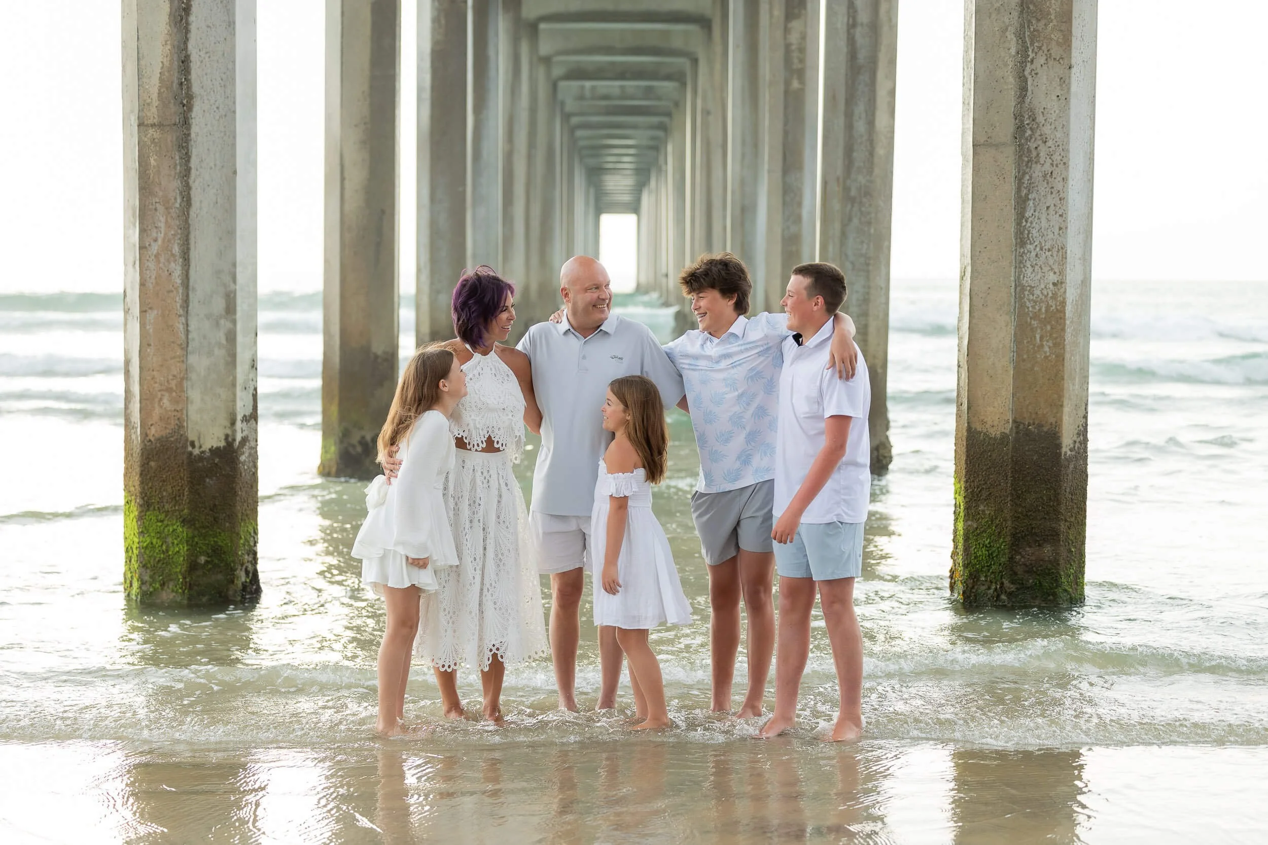 A family of seven standing in the water under a pier at the beach, smiling and embracing.