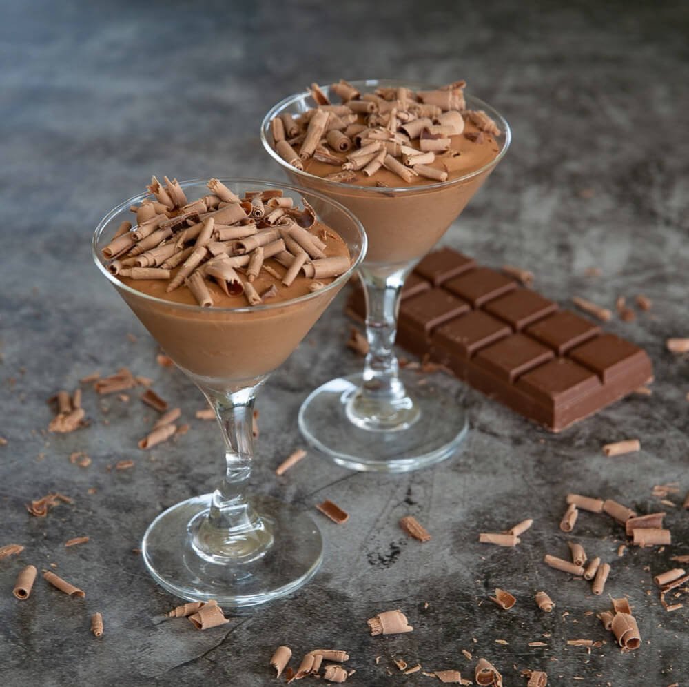 Two chocolate mousse desserts in martini glasses topped with chocolate shavings, with a chocolate bar and scattered chocolate shavings on a dark countertop.