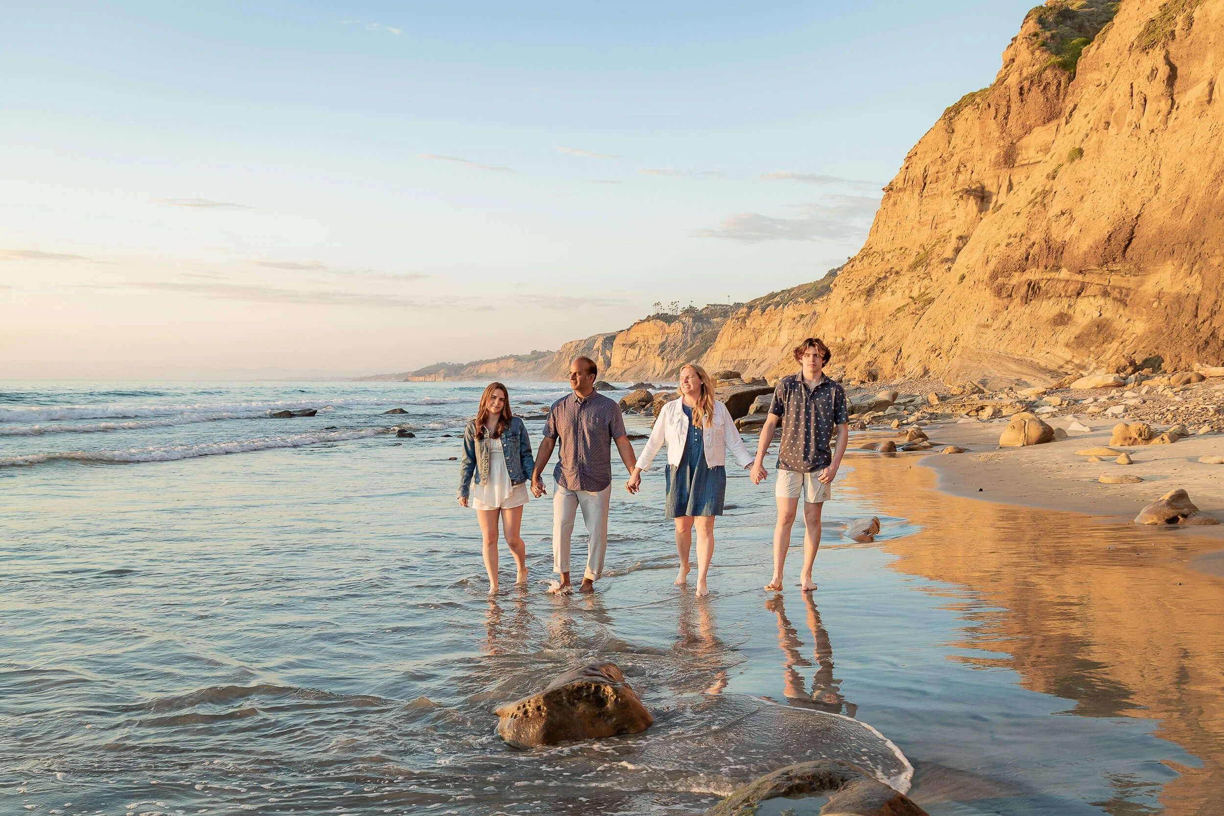 A group of four friends walking hand in hand along the beach at sunset, with cliffs in the background and waves gently crashing on the shore.